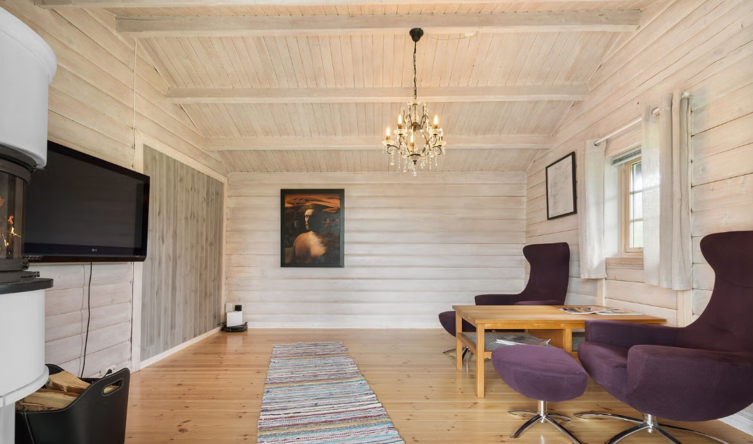 A cozy living room with light wood paneling on the walls and ceiling, a chandelier hanging in the center, a flat-screen TV mounted on the wall, two purple wingback chairs with an ottoman, a wooden coffee table, a colorful striped rug, a framed picture, and a window with white curtains.