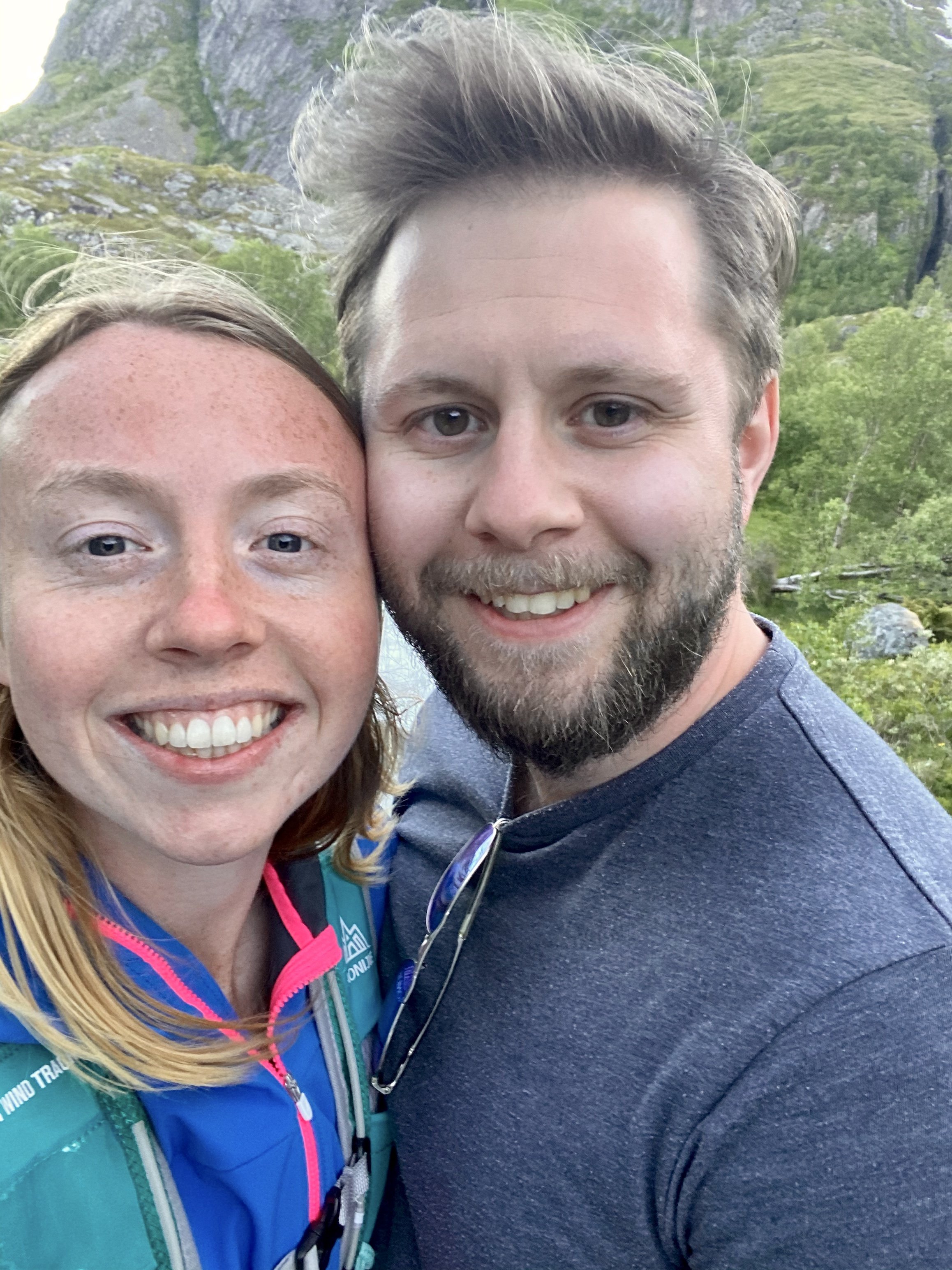 Close-up selfie of a smiling woman and a man with a scenic outdoor background of mountains and greenery.