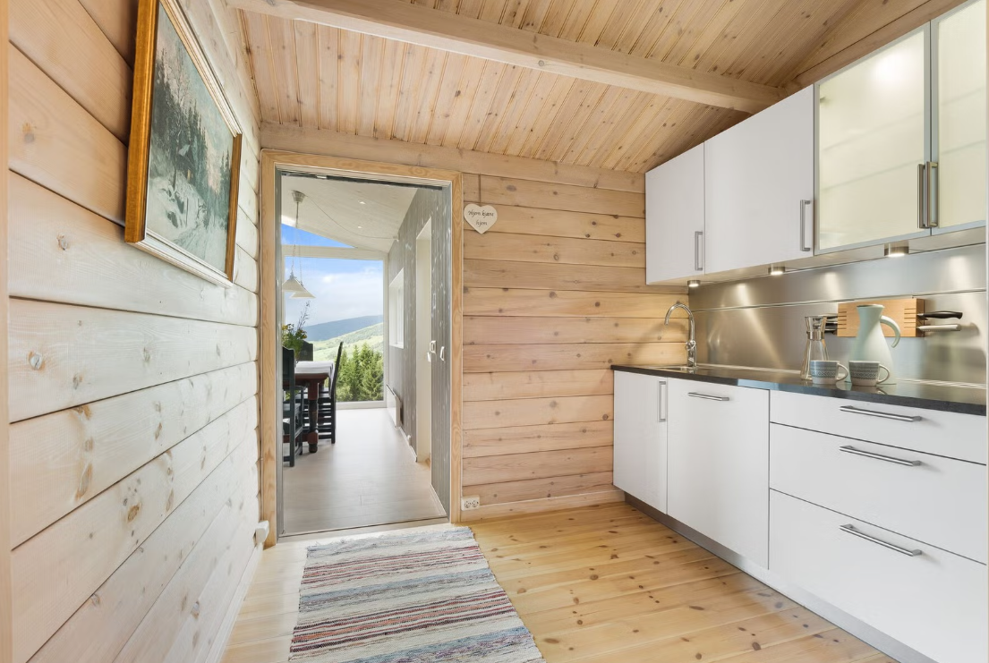 A modern kitchen with white cabinets, a stainless steel backsplash, and a wooden ceiling and floor, leading to a dining area with a view of green hills through the glass door.