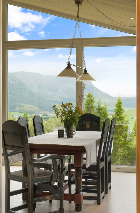 A dining table with eight chairs and a flower arrangement in a sunroom with large windows showing a mountain and valley landscape.