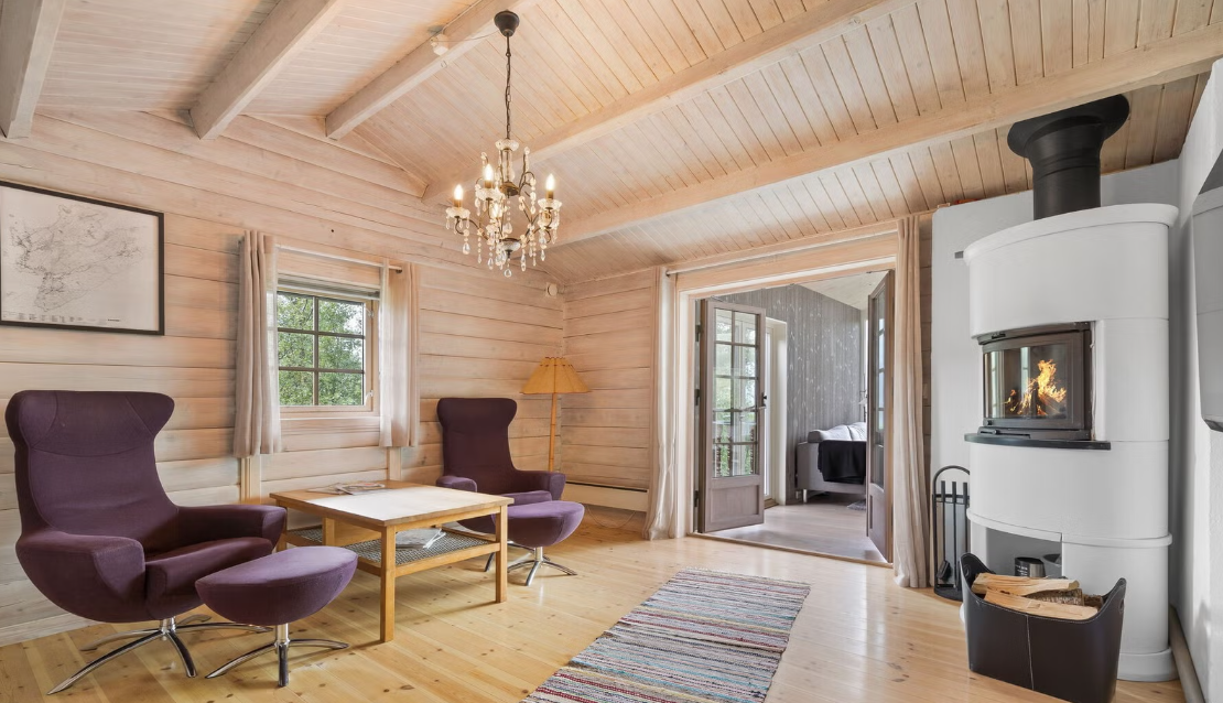 Living room with wood-paneled walls and ceiling, two purple armchairs, a wooden coffee table, a colorful rug, a fireplace with a fire, a chandelier, side table with lamp, and a door leading to another room.