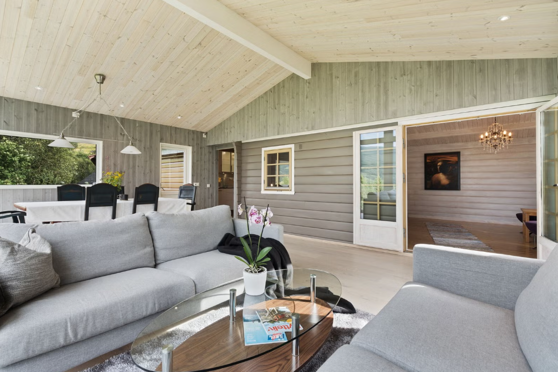 Bright living room with light wood vaulted ceiling, gray walls, and large windows opening to greenery, furniture includes a gray sofa, armchair, glass coffee table, and a dining table with black chairs, with plants and artwork visible.
