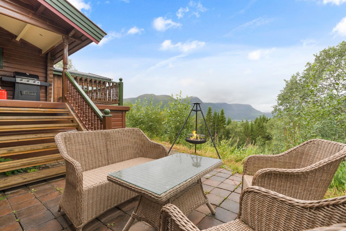 Outdoor patio with wicker furniture, a glass-top table, and a grill on the steps of a wooden house, overlooking a green forest with mountains in the background under a partly cloudy sky.
