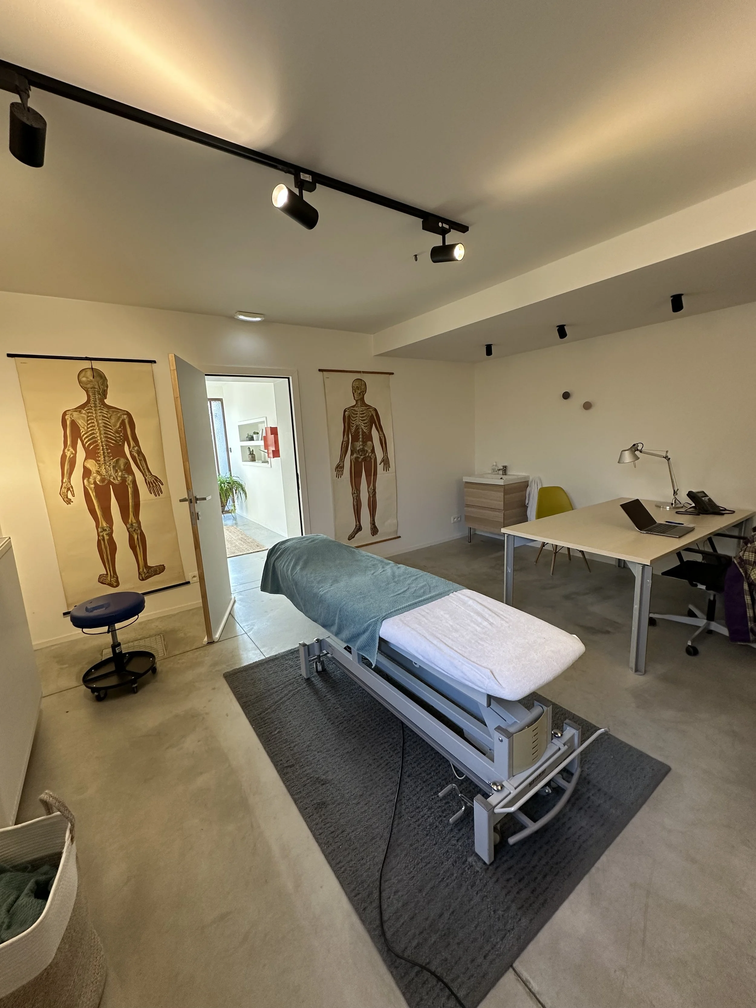 Medical examination room with an examination bed, two posters of human skeletal systems, a desk with a laptop, telephone, and lamp, and an open door leading to another room.