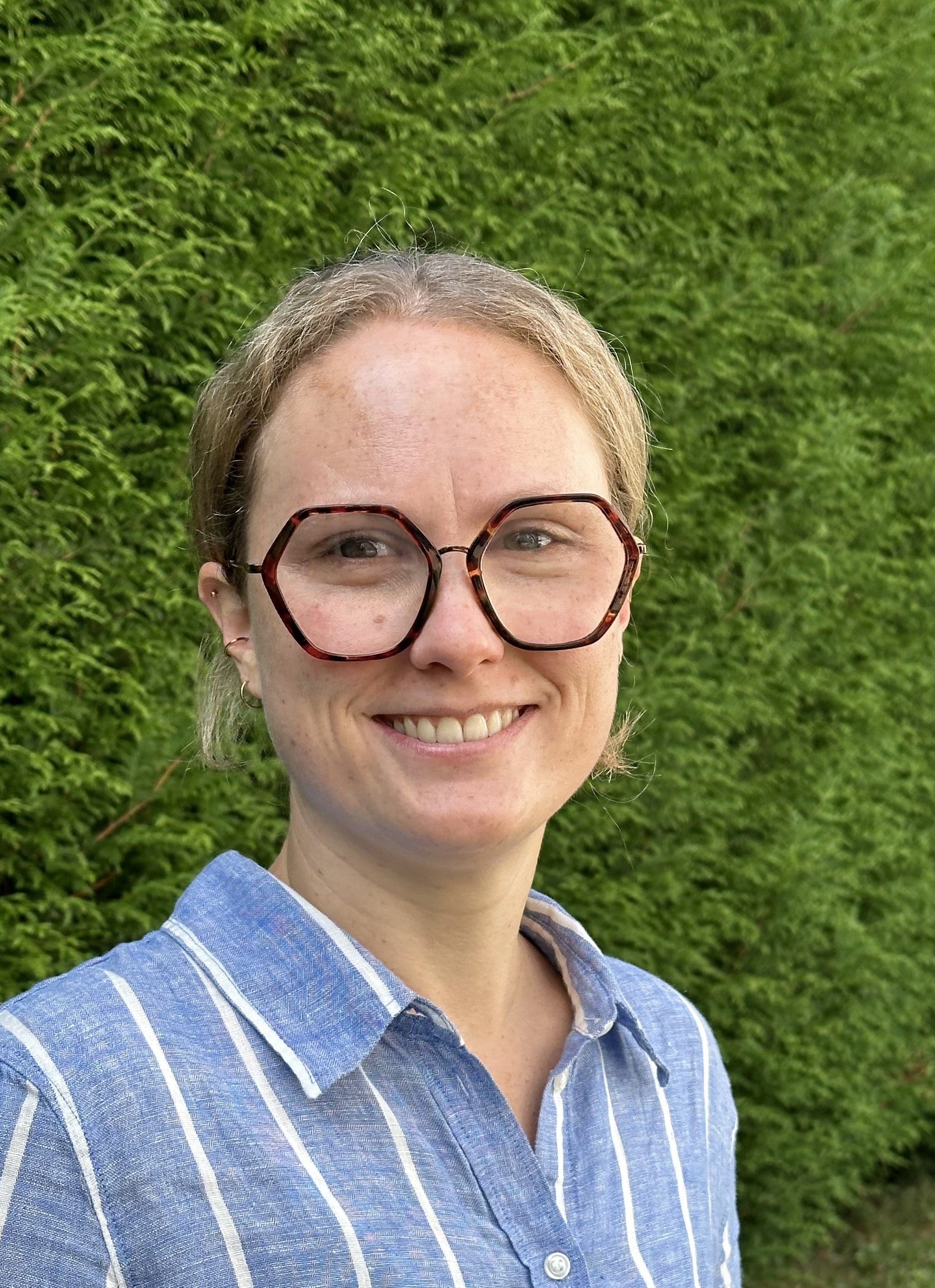 Close-up of a woman with short blonde hair wearing large tortoiseshell glasses and a blue and white striped shirt, smiling outdoors with a green hedge background.