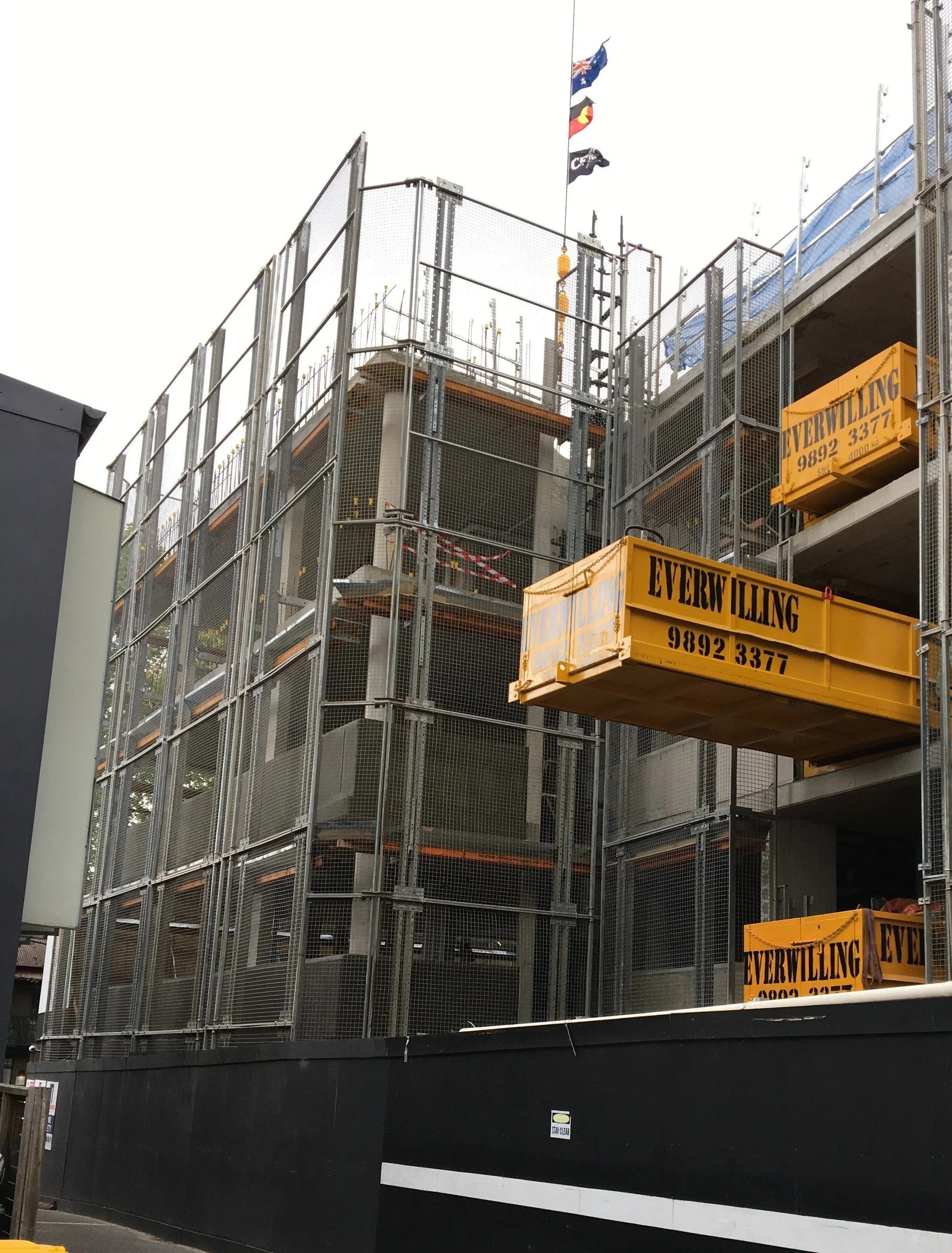 Construction site with a multi-story building under construction, surrounded by safety fences, with flags on top and yellow construction containers labeled 'Everwilling'.