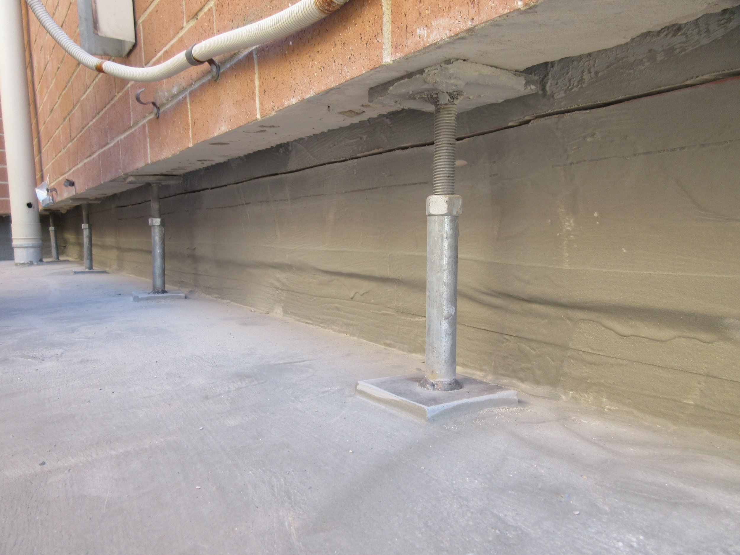 Under-construction area with support poles, concrete ground, and unfinished building structure with exposed pipes and brick wall.
