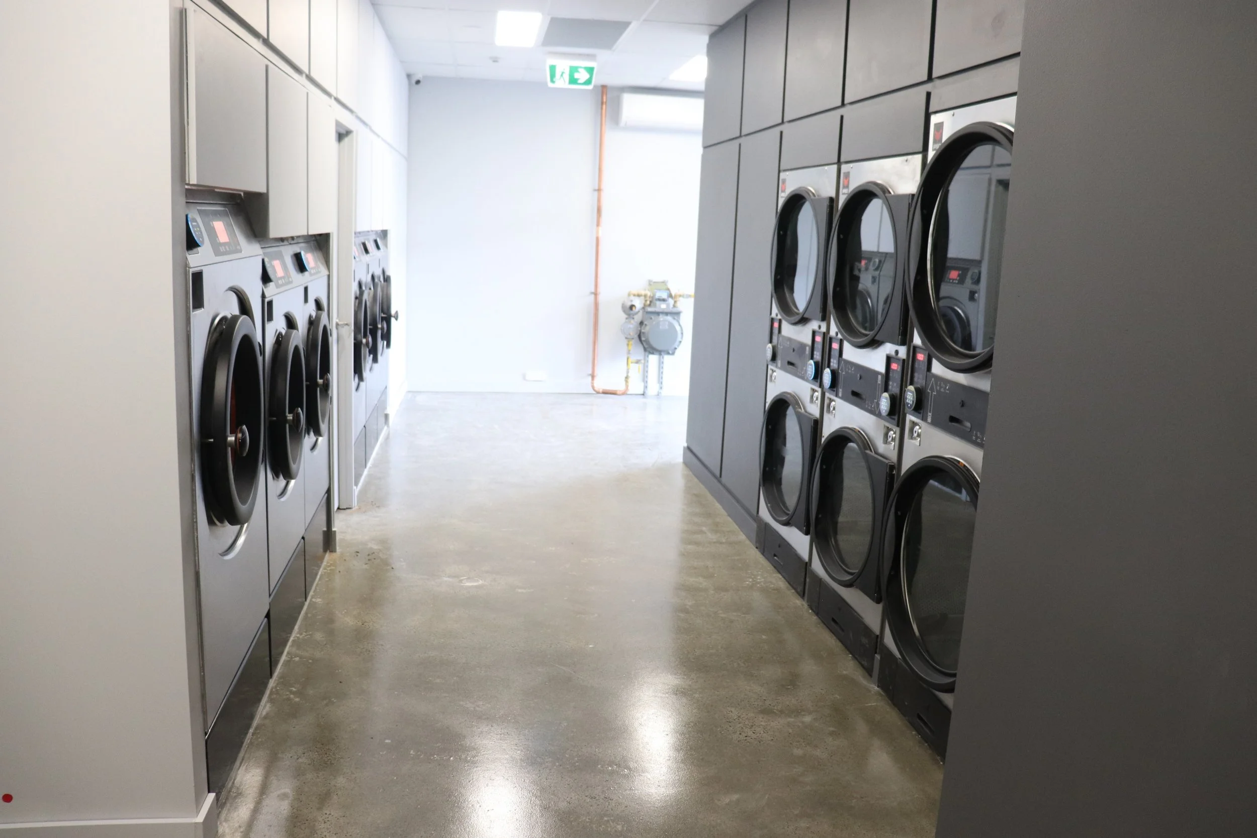 A laundromat with washing machines on both sides of a narrow corridor with a concrete floor, white walls, ceiling lights, and an emergency exit sign hanging from the ceiling.