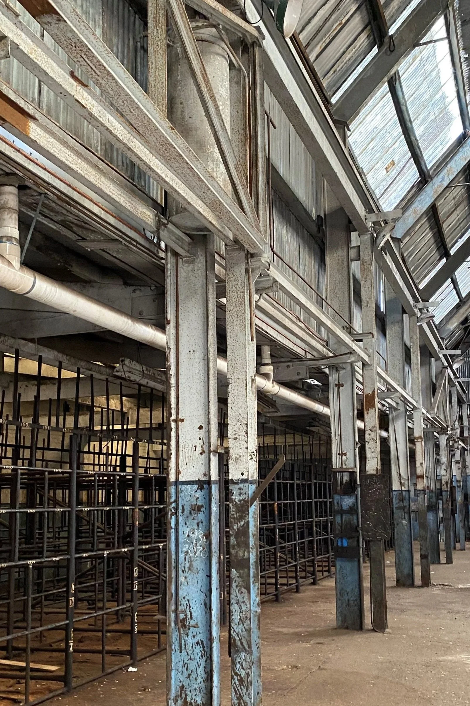 Interior of a building with exposed metal beams, rusty pipes, and metal scaffolding or framework. The roof is made of translucent panels allowing natural light inside.