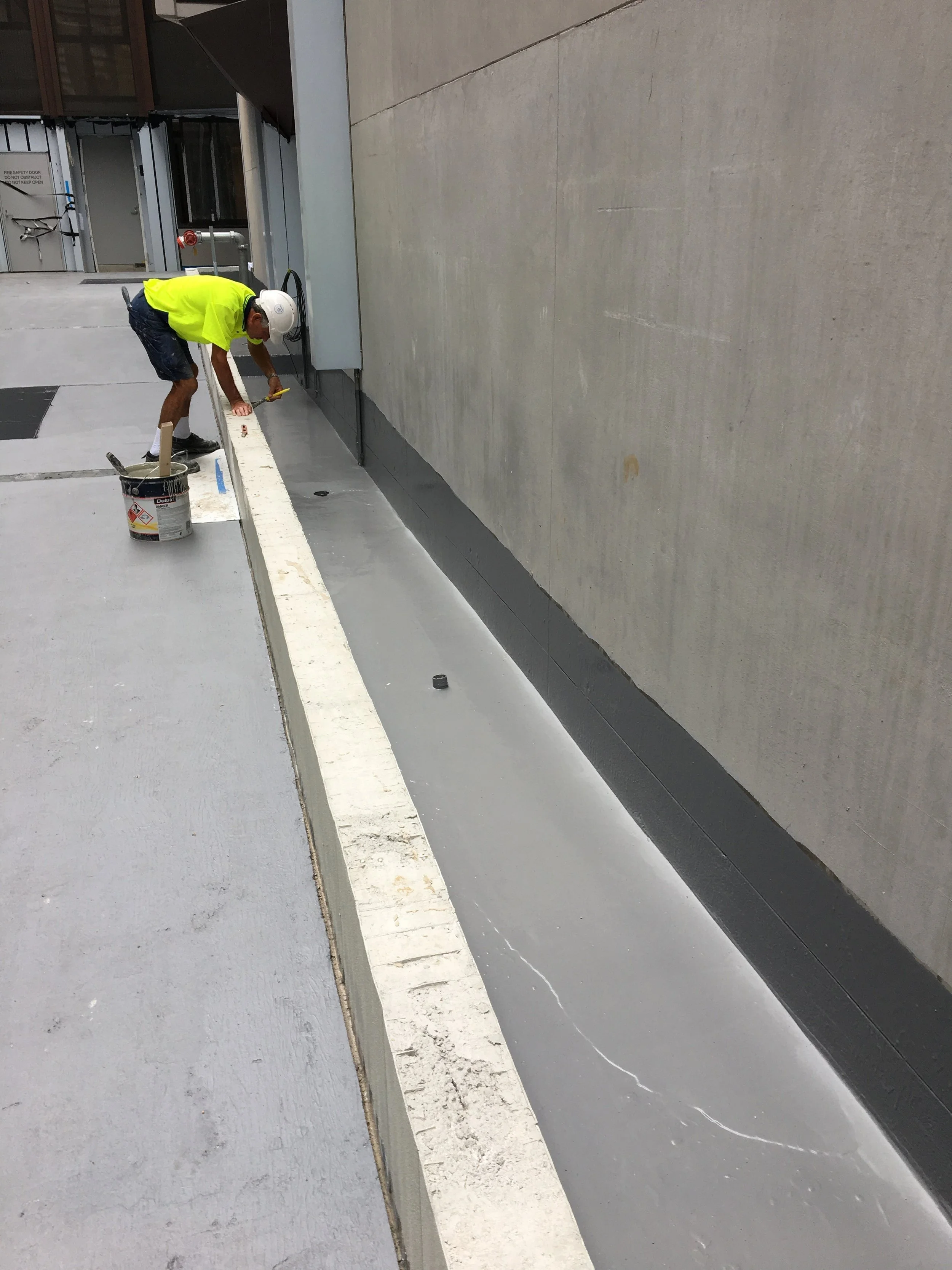 Construction worker wearing a white hard hat and a bright yellow shirt applying sealant or waterproofing to a concrete or asphalt surface along the edge of a building wall.