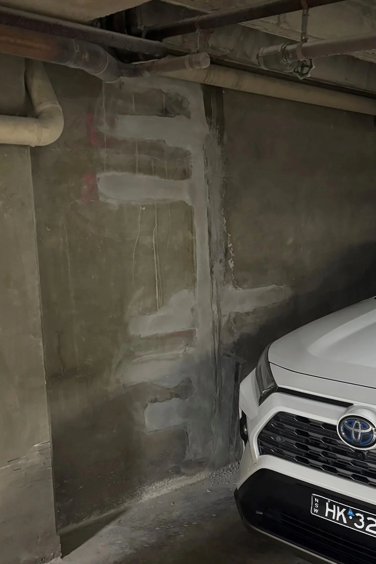 A white car parked in a garage next to a wall with exposed pipes and unfinished concrete.