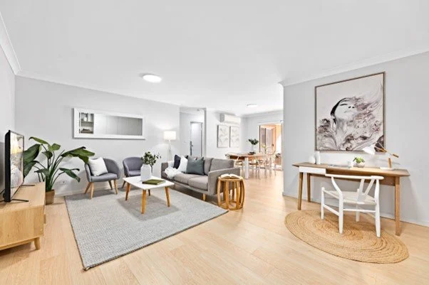 Bright living room with white walls, wooden floors, and modern furniture including a sofa, armchairs, a coffee table, and a desk with a chair. Decor includes artwork, plants, and a rug.