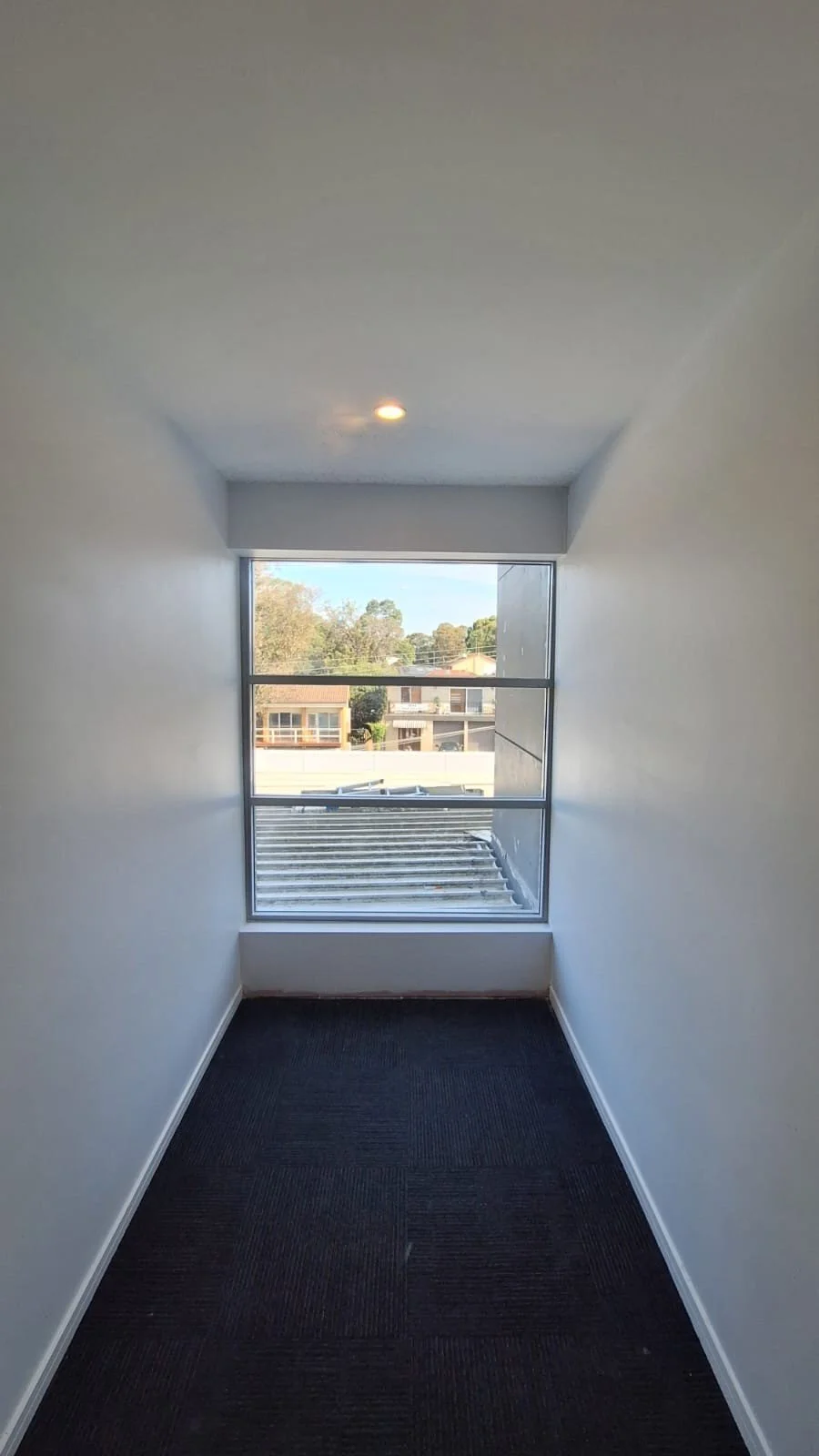 Empty narrow hallway with white walls, black carpet flooring, and a large window at the end showing an exterior view of trees and buildings.