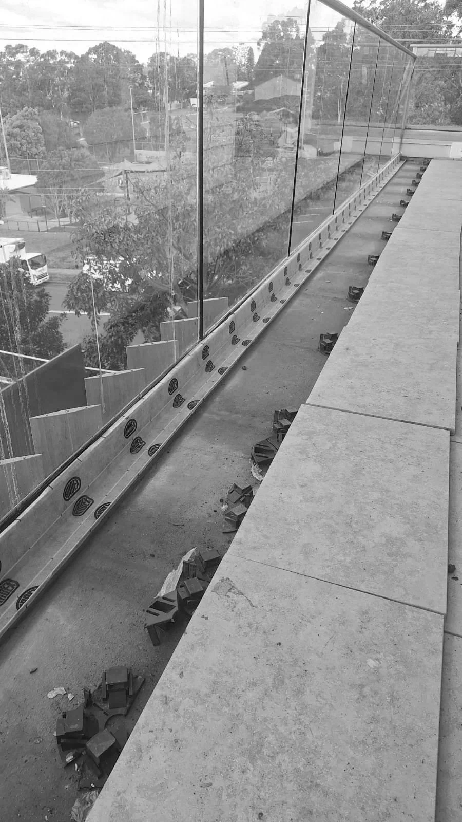 Construction site on a balcony or rooftop with glass railings and concrete flooring. Building materials and tools are scattered on the floor, with a view of trees and houses in the background.
