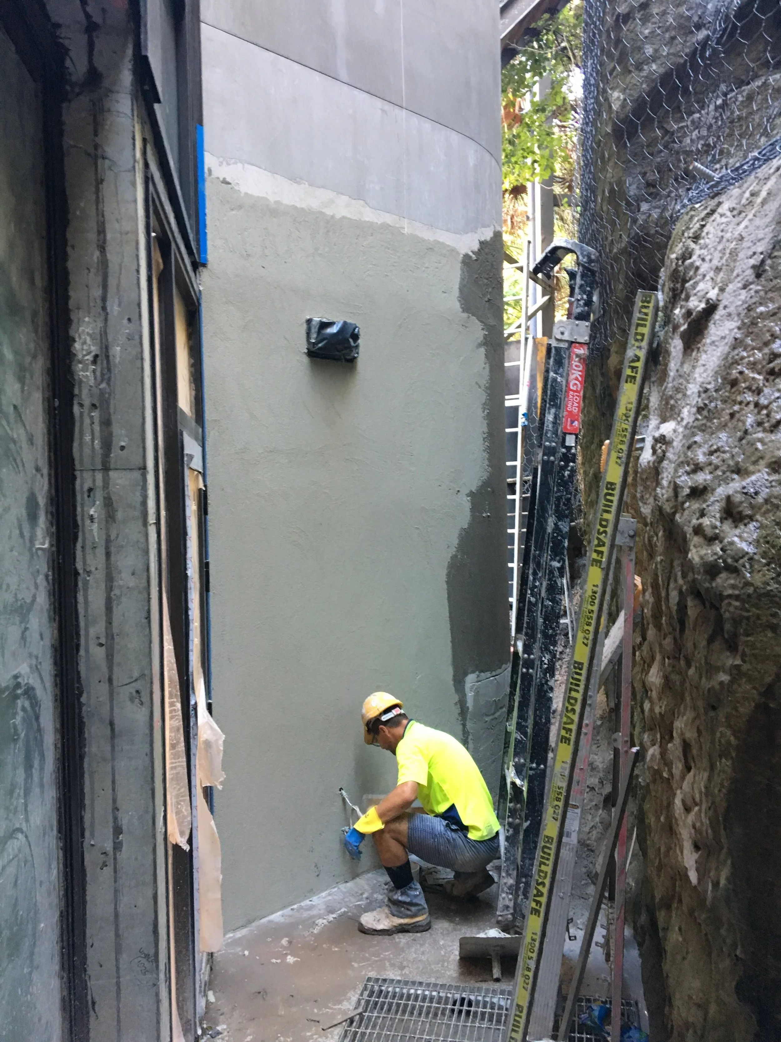 Construction worker wearing a yellow helment and bright yellow shirt working on cement wall at a construction site with scaffolding and safety tape.