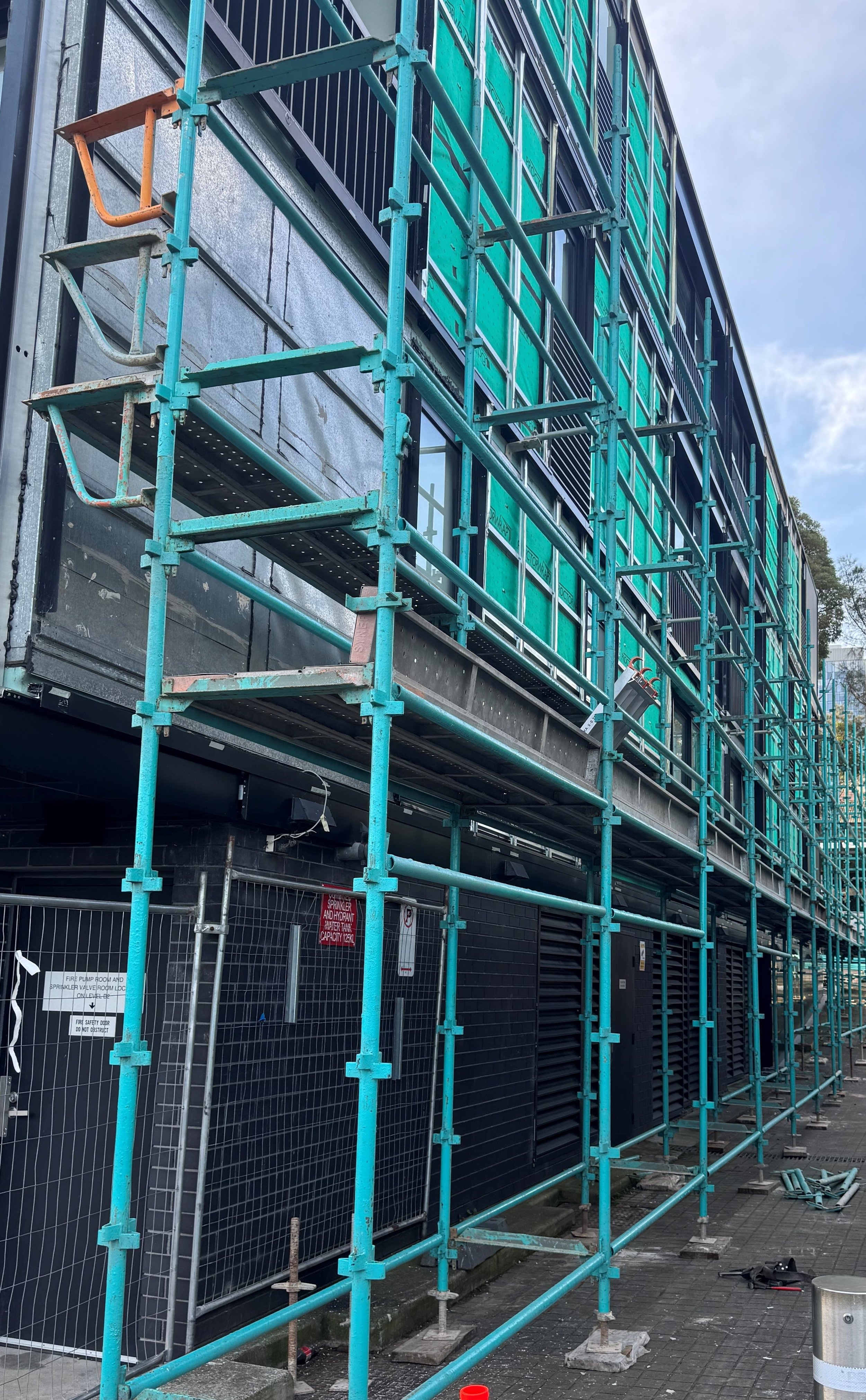 Construction site with teal metal scaffolding in front of a black building with windows. Crates and signs are visible behind a safety fence at street level.