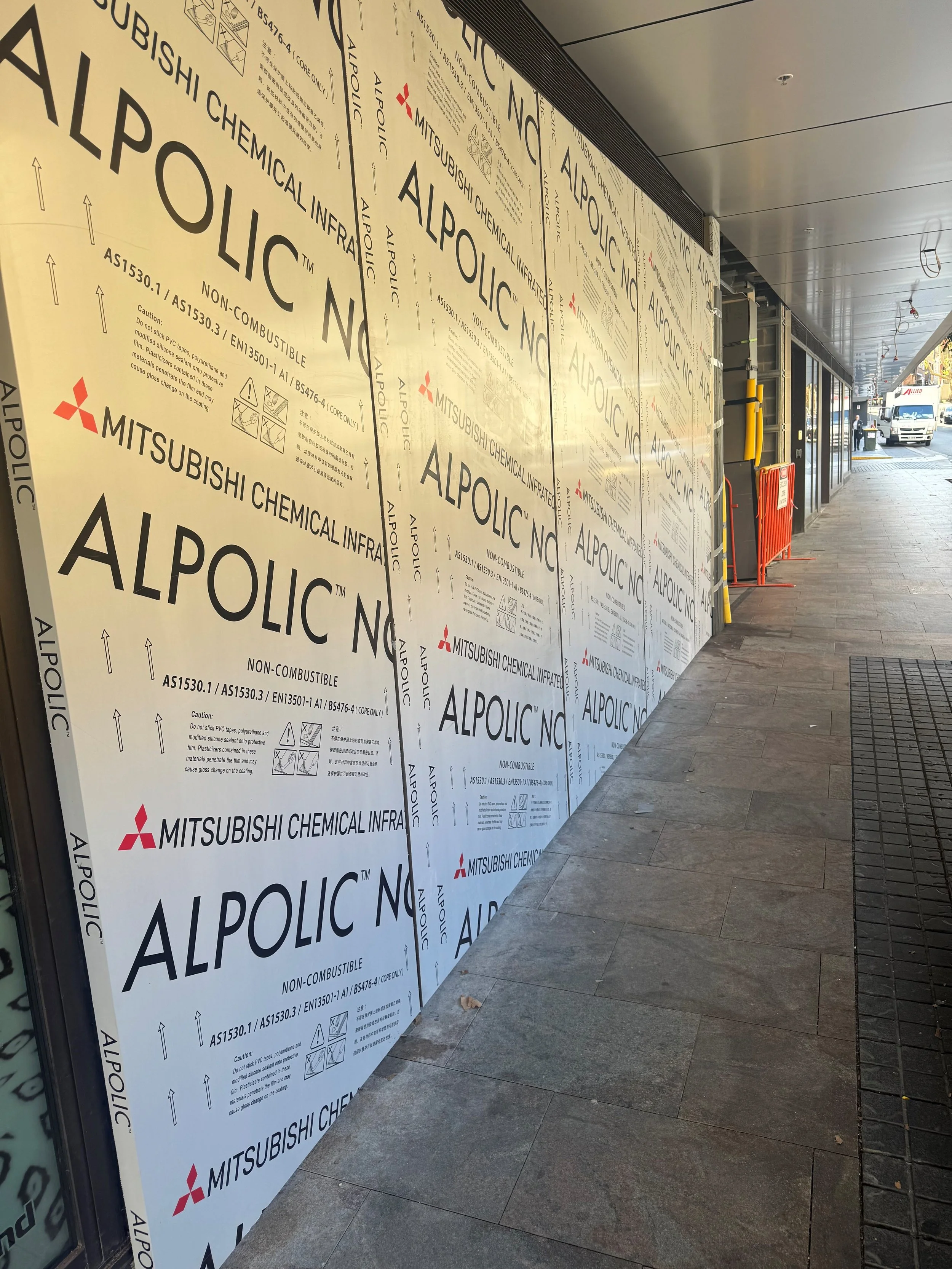 Construction area with large white panels printed with 'ALPOLIC' and warning symbols, outside a building with a sidewalk. Visible vehicles and a person in the background.