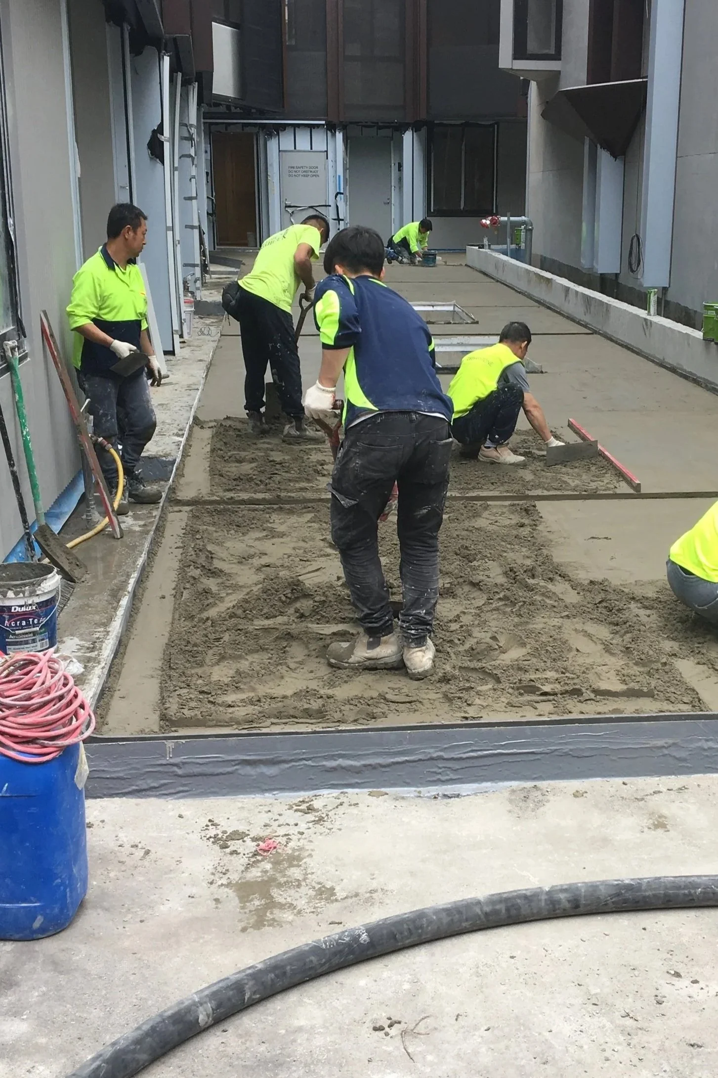 Workers in high-visibility vests and work clothes are spreading and leveling concrete in a construction area, with some crouching and others standing, and construction materials and equipment around.