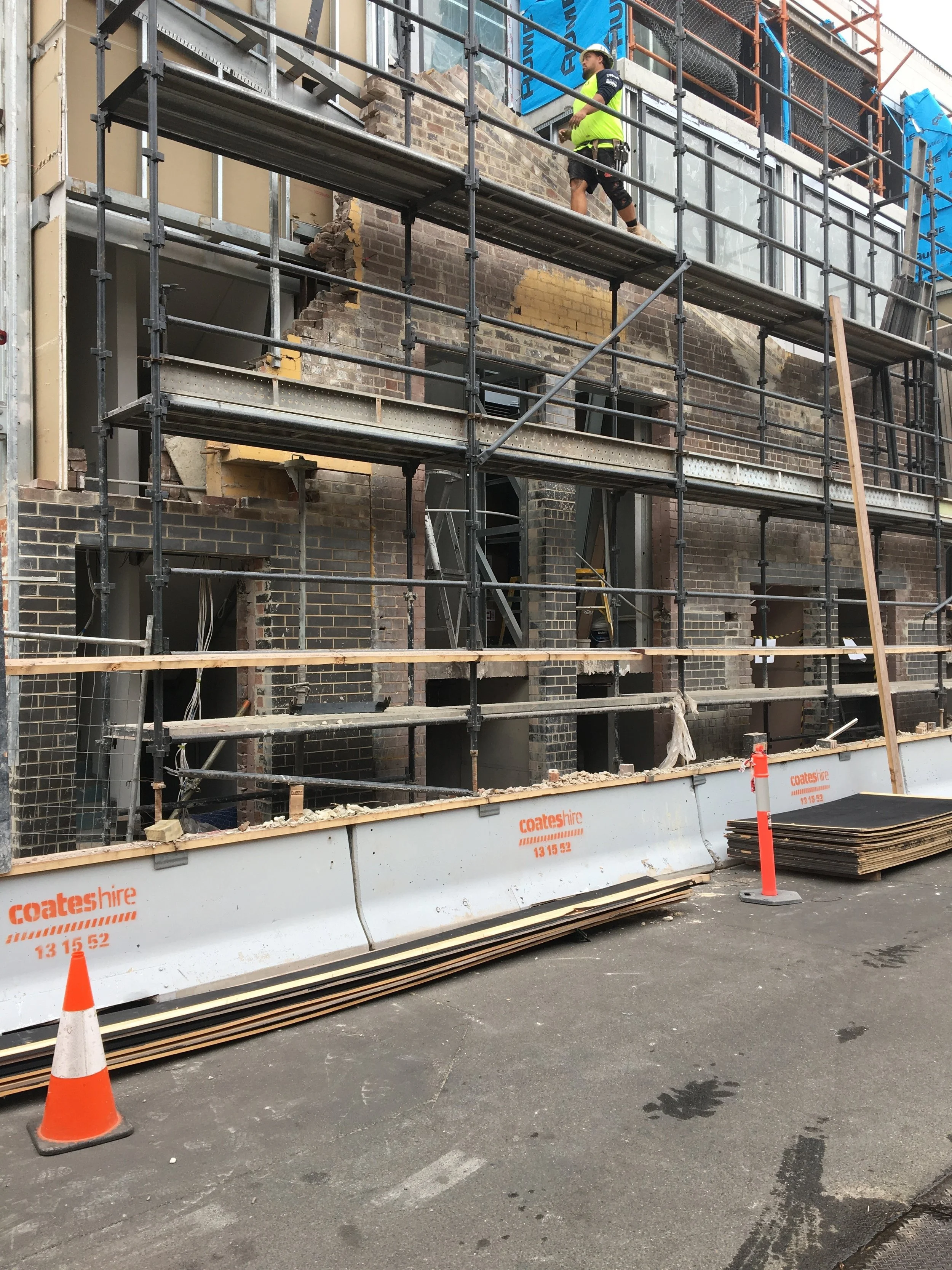 A construction site shows a building under renovation with scaffolding. A worker in a yellow safety vest, helmet, and shorts is on the scaffolding. In the foreground, there is a construction boundary with orange and white cones and a pile of wooden planks.