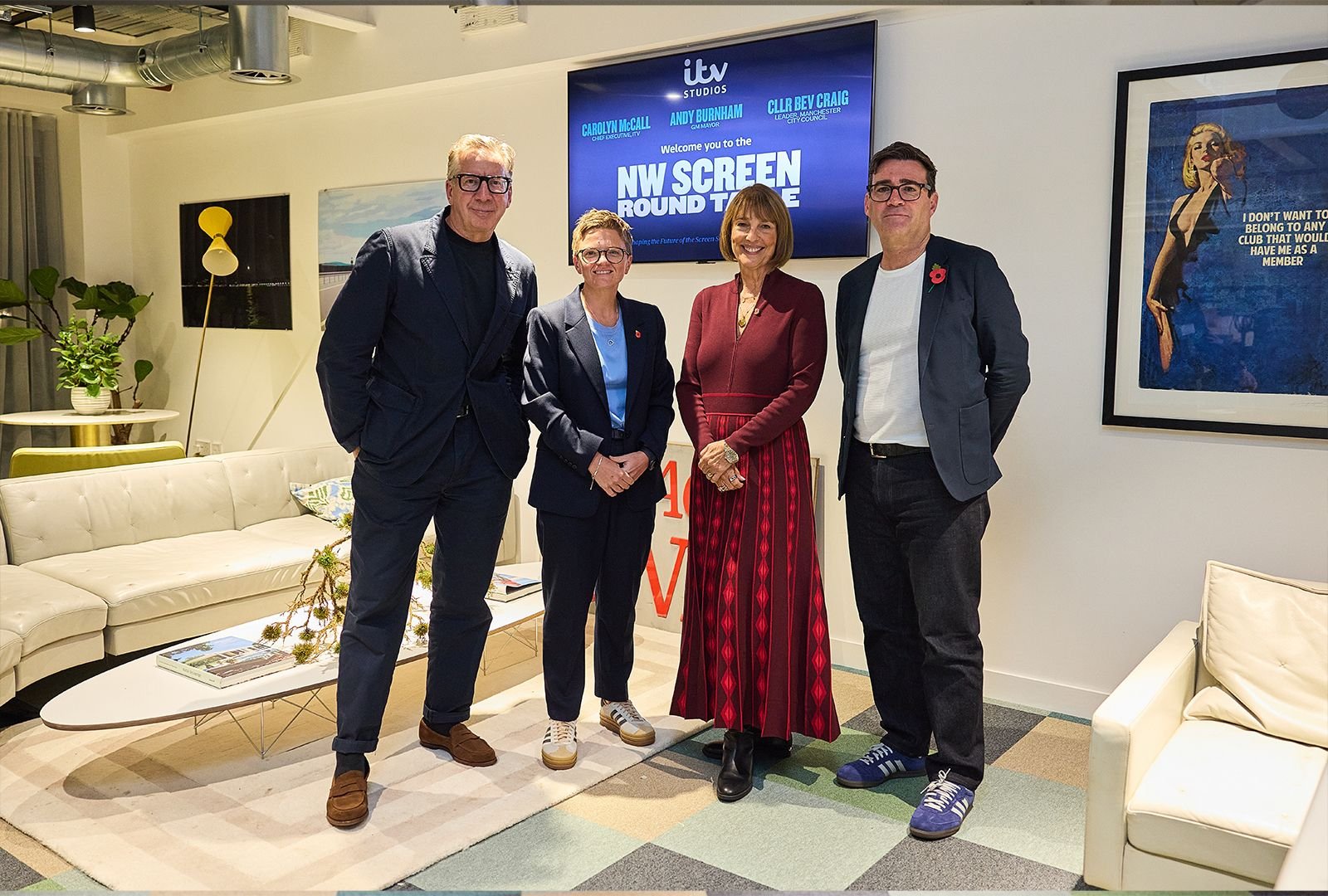 Four people standing in a modern living room with a beige couch, green plants, and wall art, posing for a photo. A large screen behind them displays the text "NW Screen Round Table" and the names of individuals.