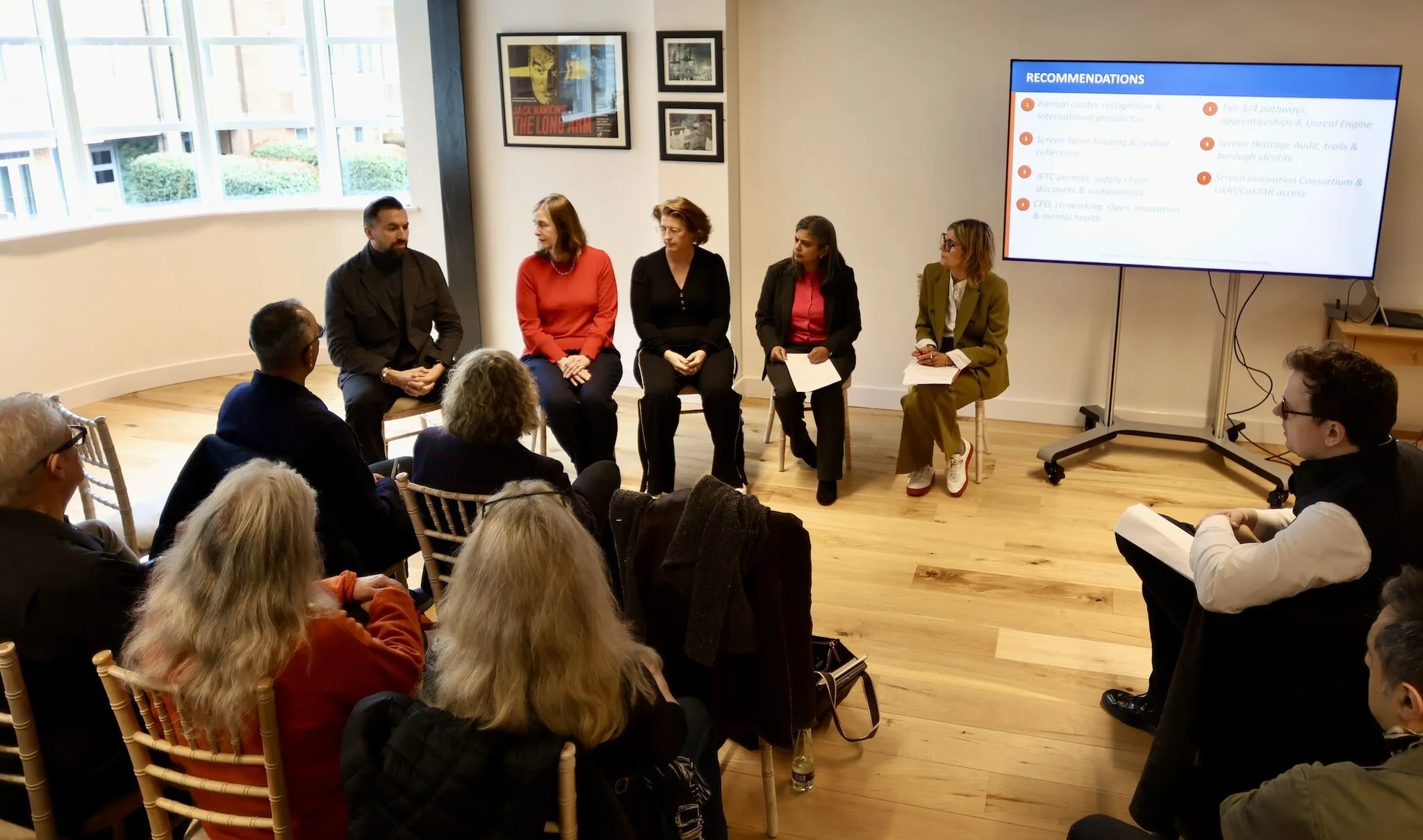 A group of six people sitting in chairs on stage during a panel discussion or presentation, with an audience watching. There is a large screen displaying a PowerPoint slide titled 'Recommendations' with six bullet points. The room has large windows on the left side and framed artwork on the wall.