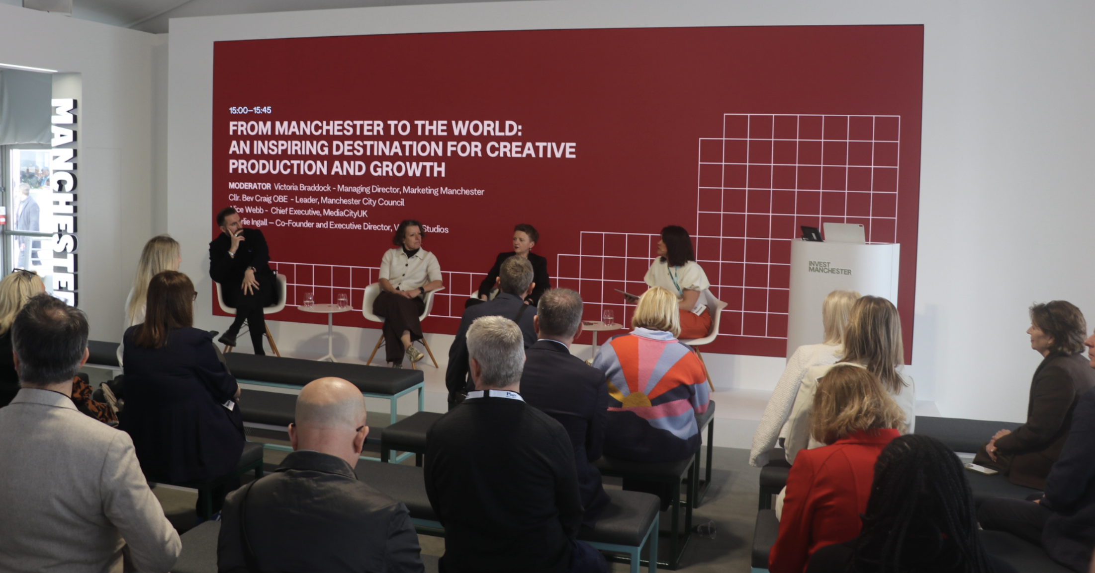 A panel discussion at a conference with four women seated on stage, audience members listening, and a large red screen behind with white text about Manchester to the world, a creative production and growth event.