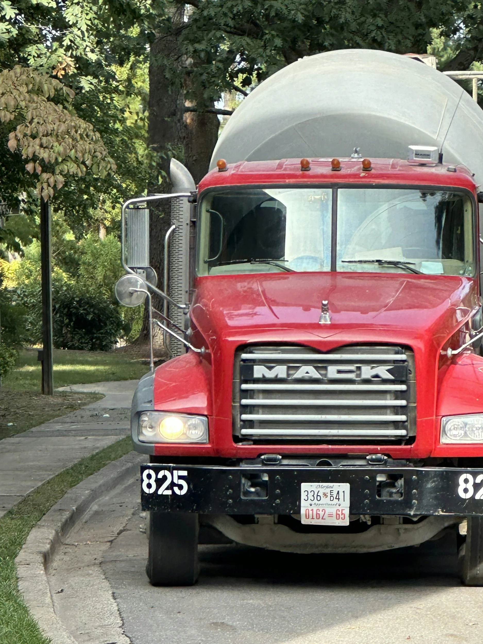 Front view of a red ready-mix Mack truck, parked on a residential street with trees and greenery in the background.