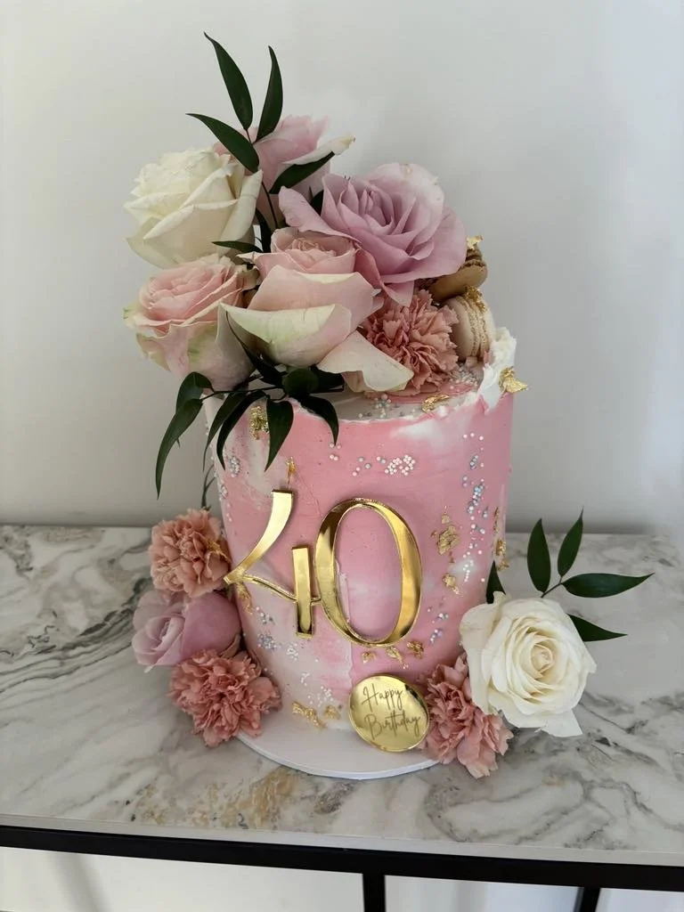 Pink birthday cake decorated with fresh roses, carnations, and greenery, with gold accents and a large gold 'KO' topper on the front, placed on a white and gray marble surface.