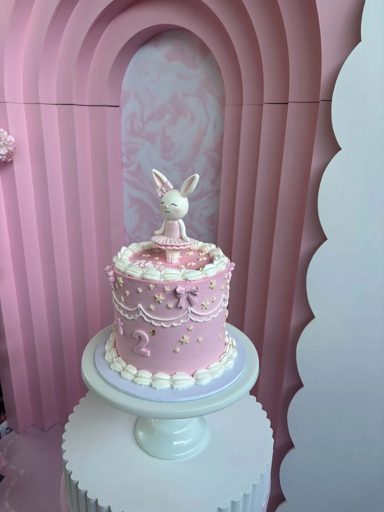 Pink birthday cake with a bunny topper, decorated with small stars and bows, on a white scalloped-edged cake stand against pink and white background.