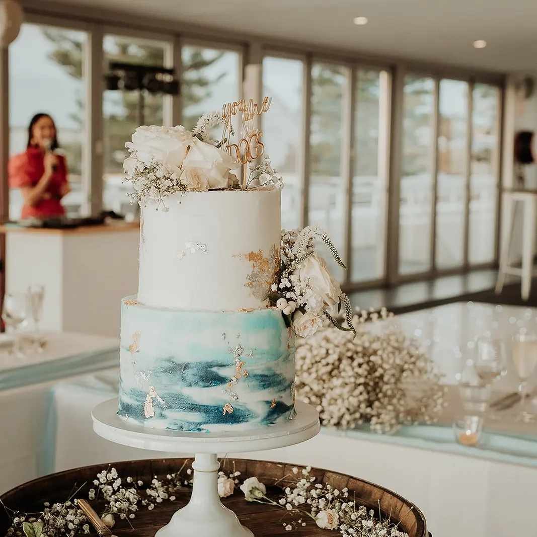 Elegant two-tiered wedding cake with white and blue marbled design, decorated with white flowers and gold accents, and a 'You are my forever' topper. The cake is displayed on a white cake stand, with a backdrop of a bright, spacious room with large windows and a woman speaking into a microphone in the background.