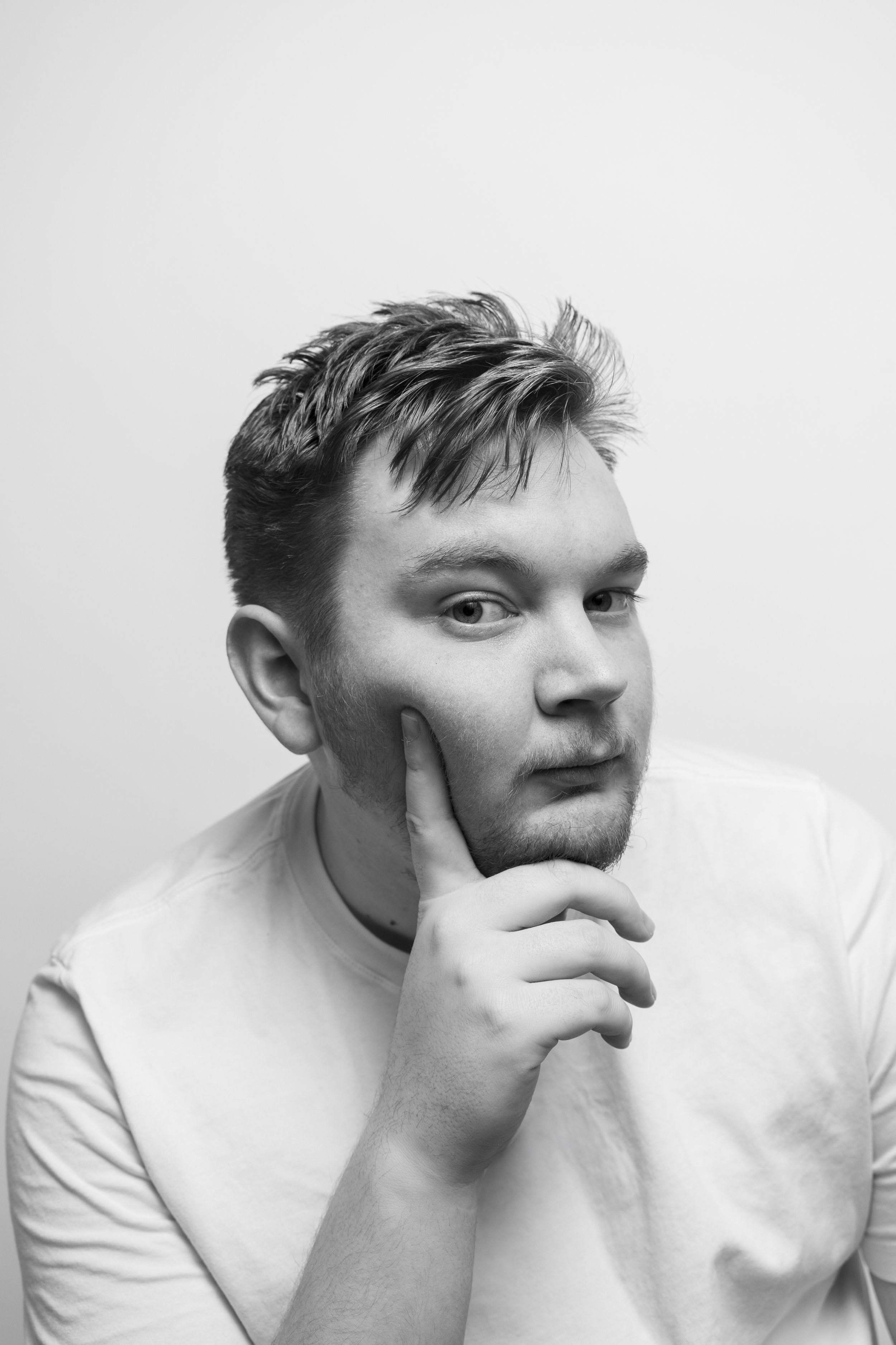 Black and white photo of a young man with styled hair, touching his chin with his right hand, wearing a light-colored t-shirt.
