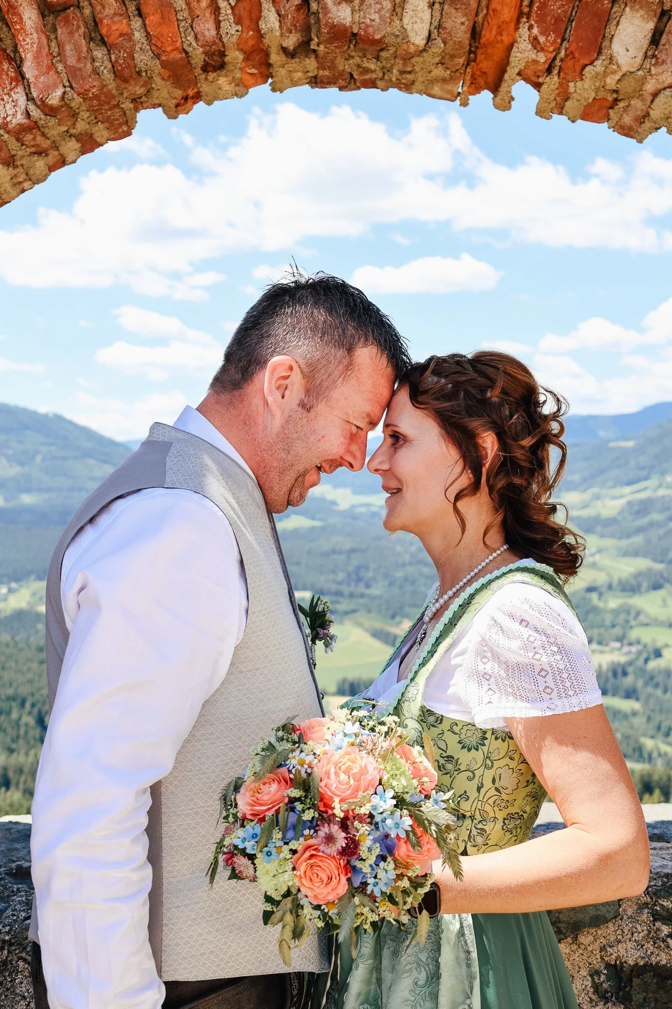 Ein Paar bei einer Hochzeit, das sich an den Stirnen berührt, vor einer Kulisse von Bergen und Himmel, mit einem Blumenstrauß in der Hand.