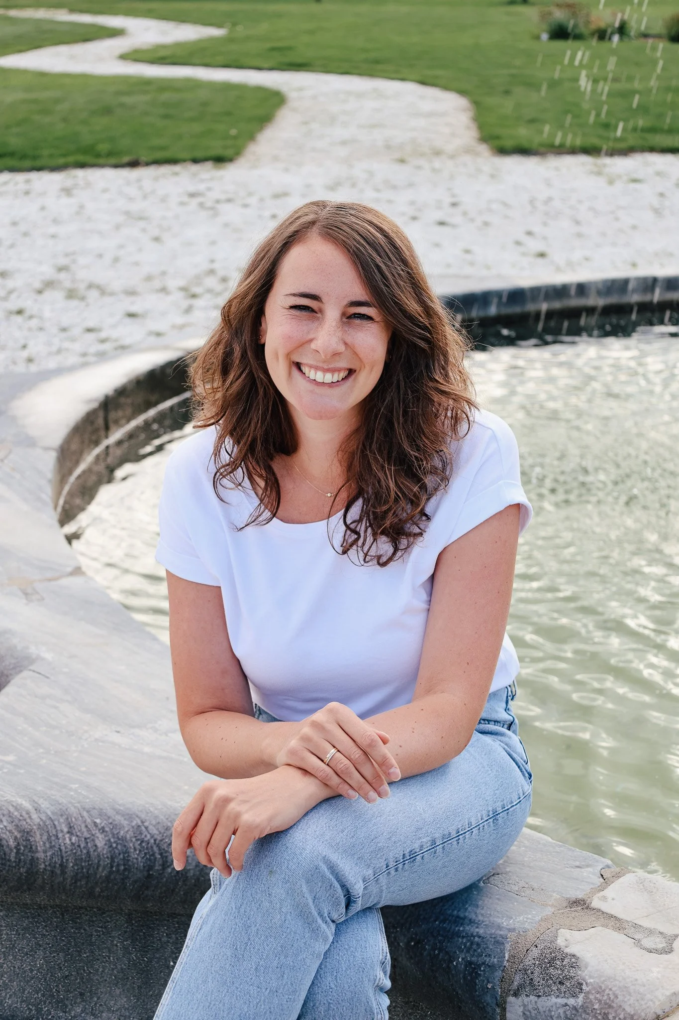 Junge Frau sitzt am Wasser, lächelt, trägt weißes T-Shirt und blaue Jeans, im Hintergrund grüne Wiesen und ein kleiner Wasserfall.