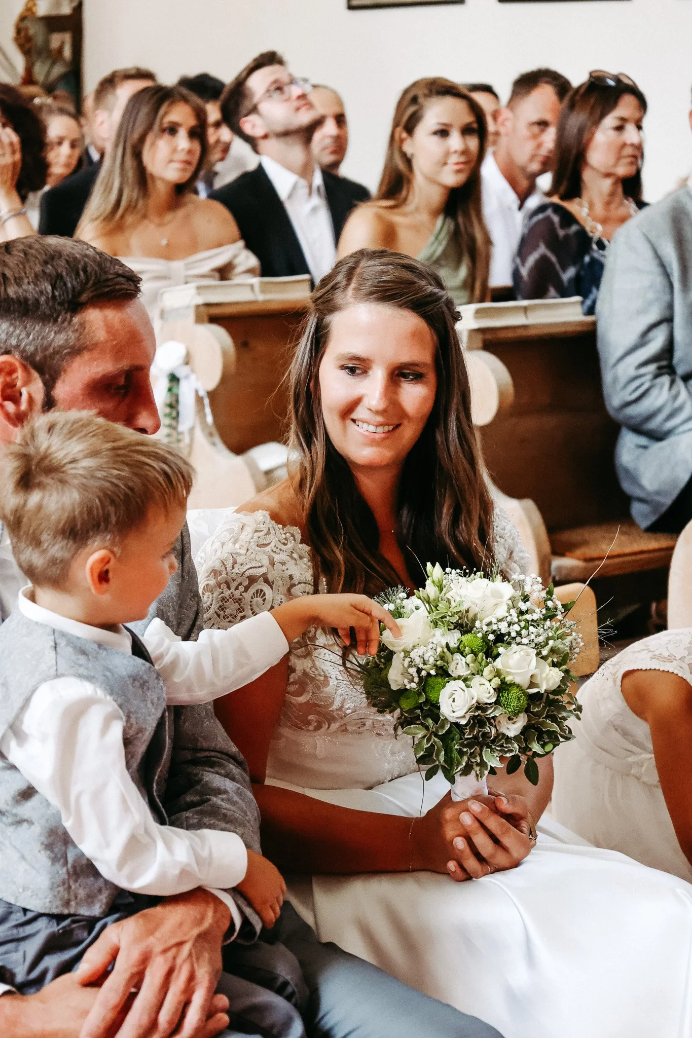 Eine Braut hält einen Blumenstrauß und ist bei einer Hochzeit, umgeben von Gästen, die im Hintergrund sitzen.