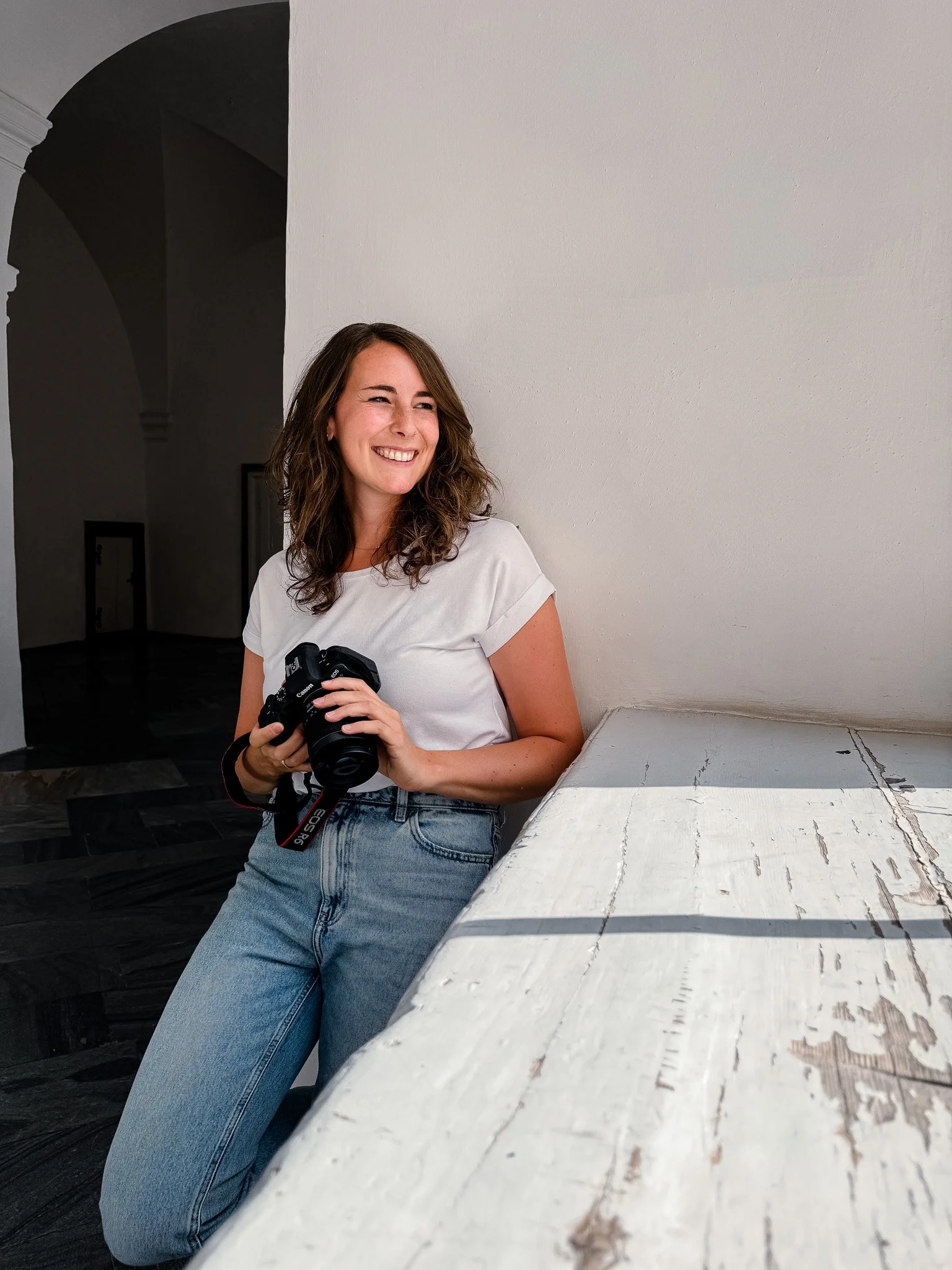 Eine lachende junge Frau mit braunen, lockigen Haaren, die eine Kamera hält, steht neben einem weißen, alten Holztisch in einem Raum mit dunklem Hintergrund und einem weißen Wand.
