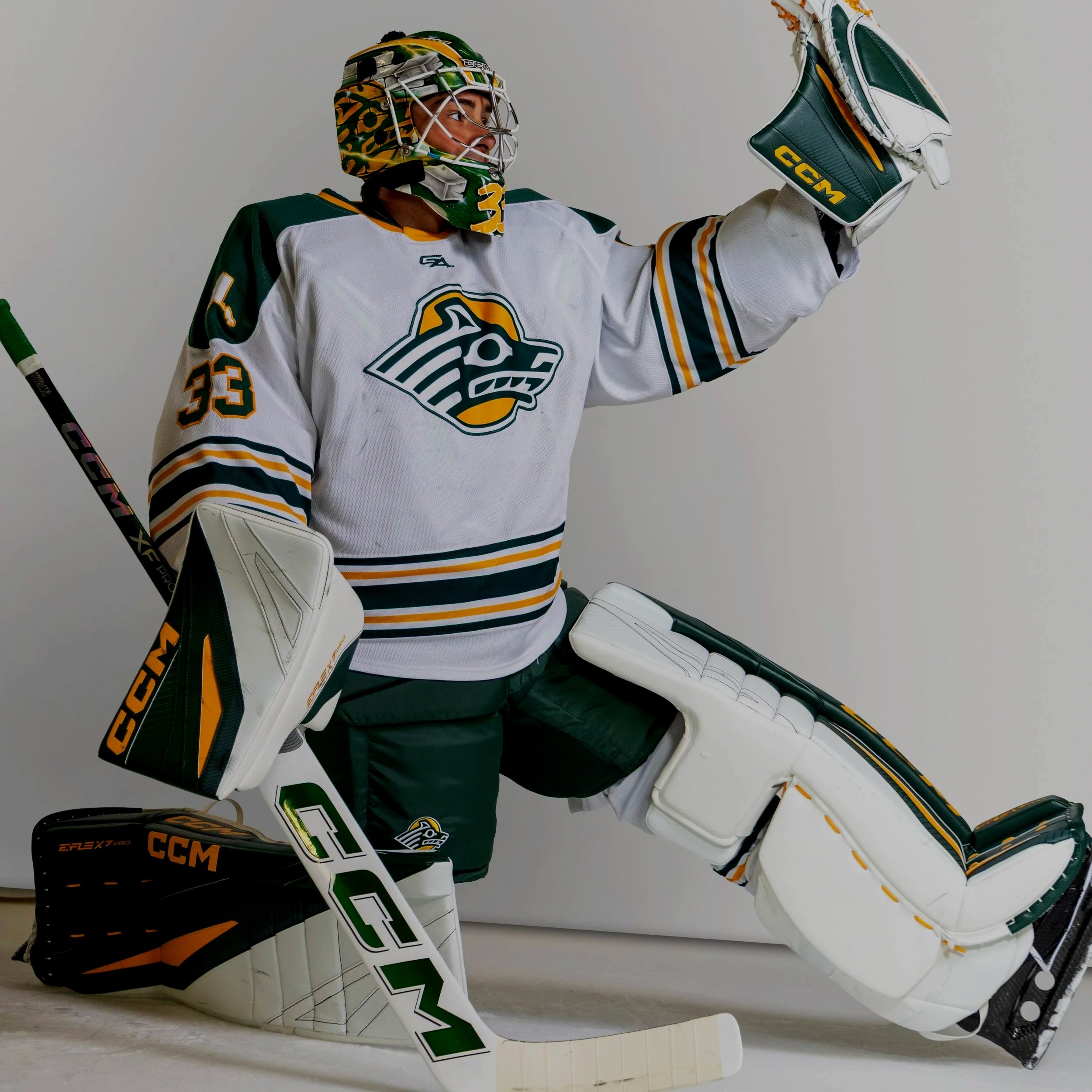 Hockey goalie in uniform with a logo on his jersey, wearing a helmet, gloves, and pads, holding a hockey stick, and standing next to his white hockey goalie skate. The background is plain and off-white.