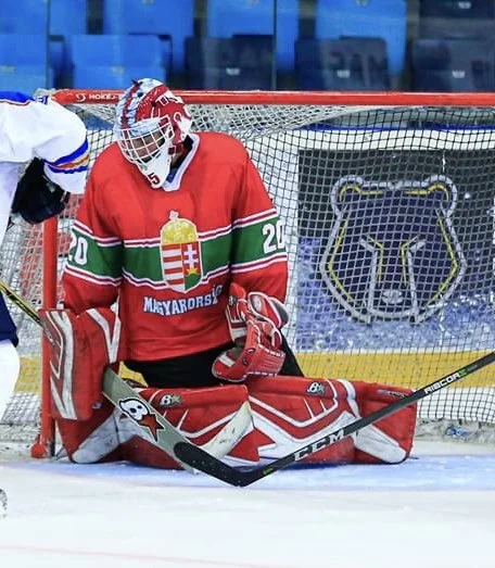 Hockey goalie in red jersey with Hungarian emblem, sitting on ice in front of the goal net.
