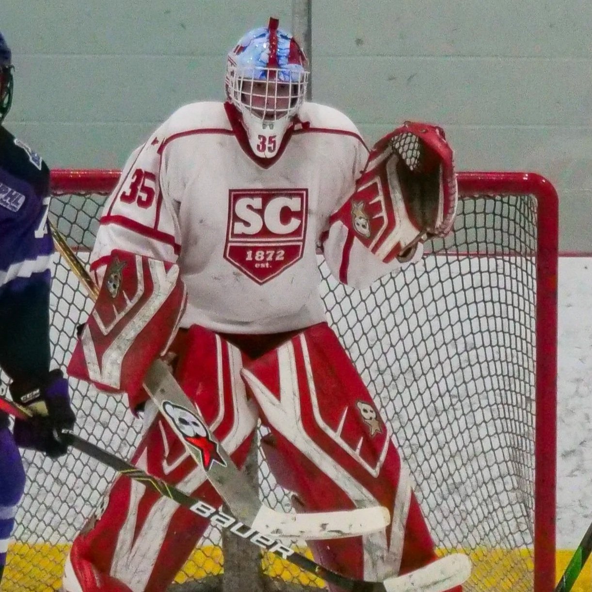 Ice hockey goalie in white and red gear standing in front of goal net on an ice rink.
