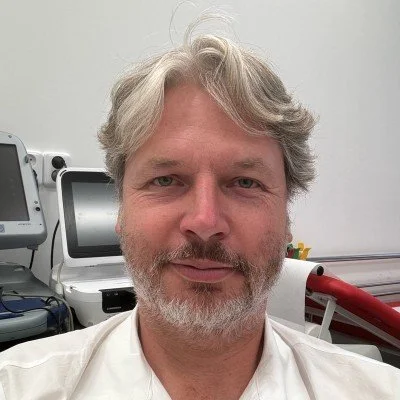 A middle-aged man with gray hair and a beard, wearing a white shirt, indoors with medical equipment in the background.