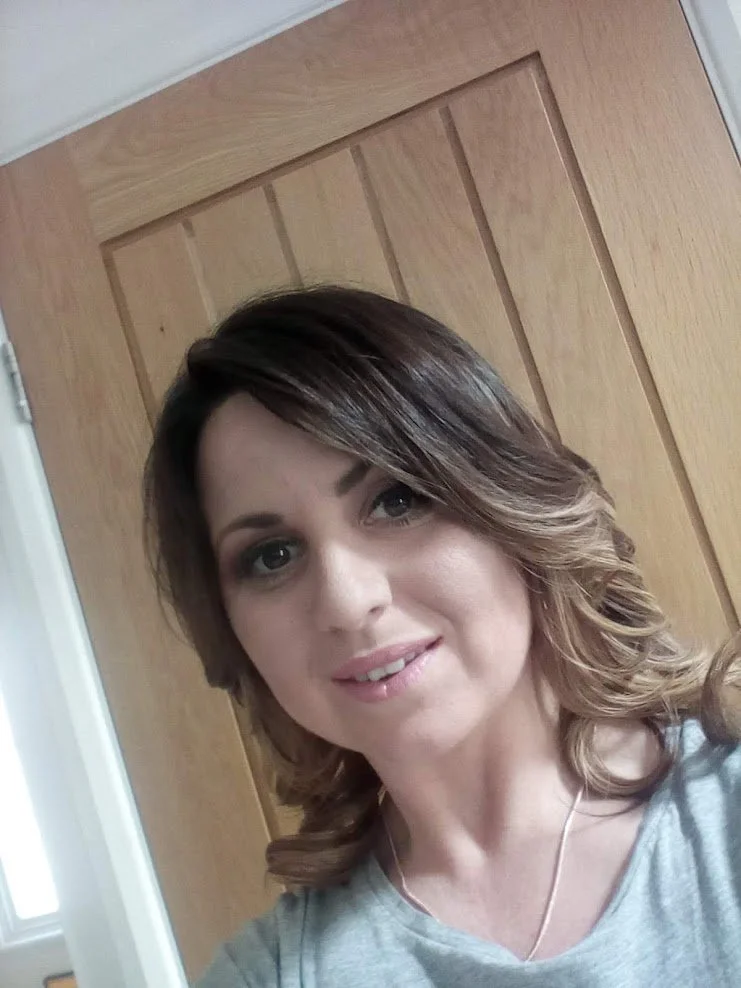 A woman with shoulder-length brown hair and a light gray shirt smiling at the camera, standing in front of a wooden door.