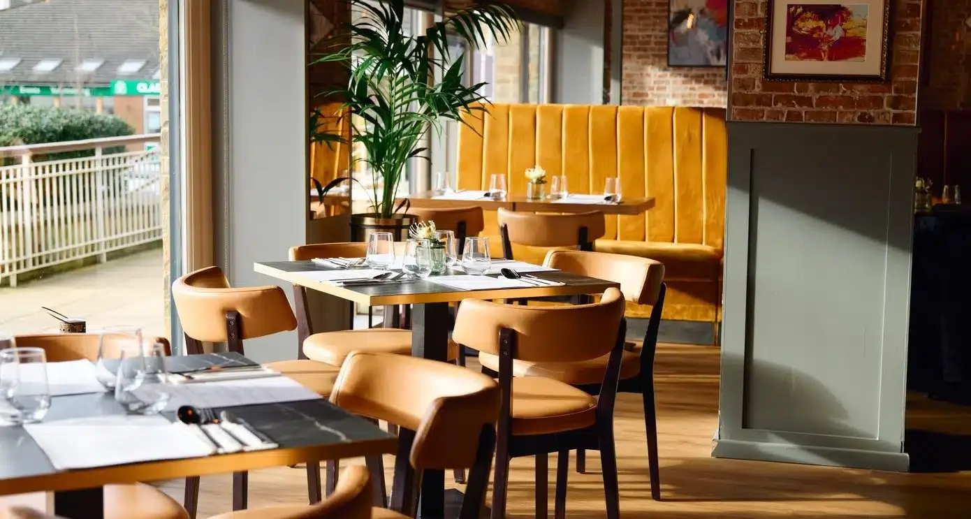 Interior of a restaurant with wooden tables, tan chairs, a yellow booth, large window, potted plant, and framed artwork on brick walls.