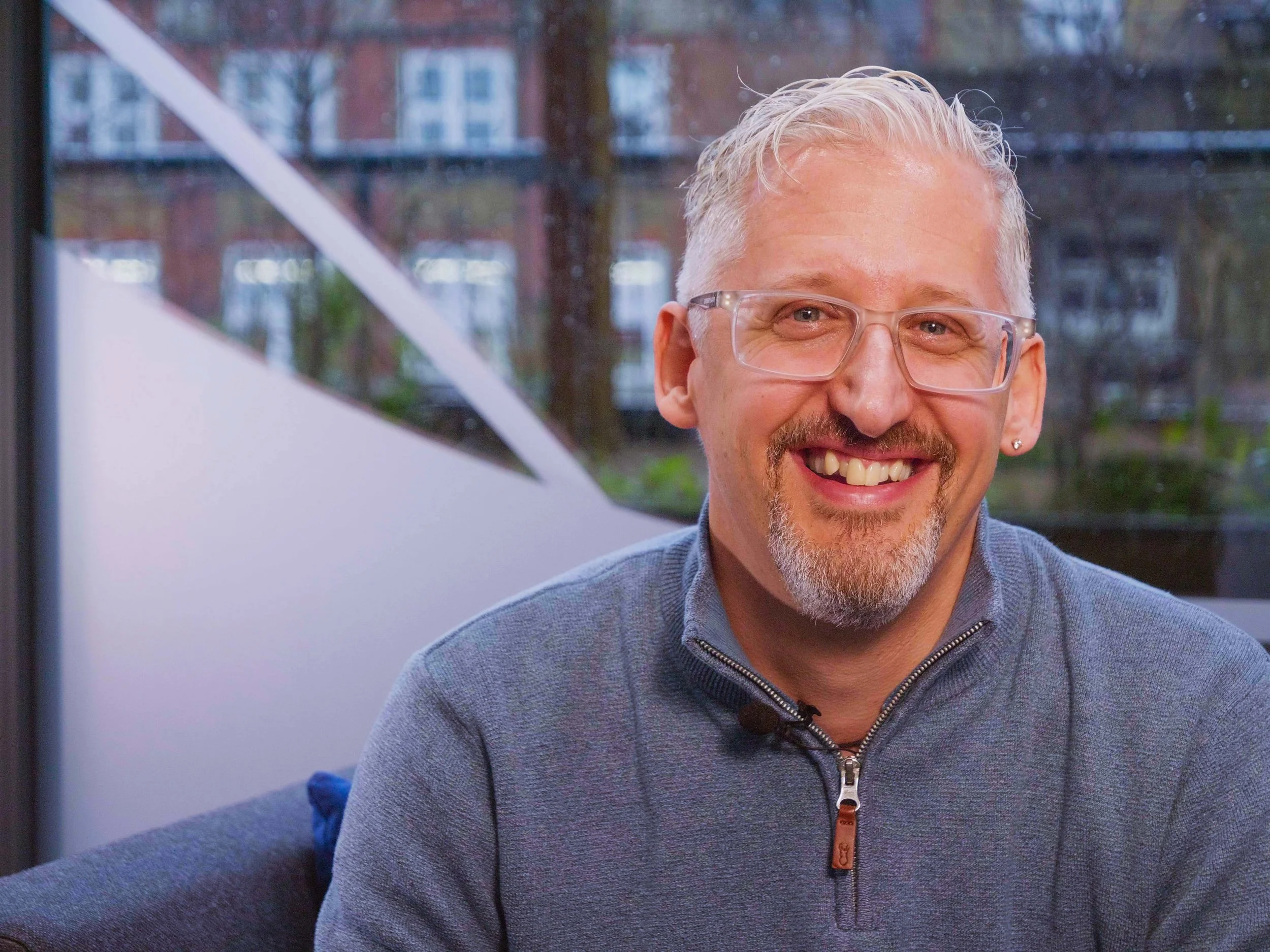 Smiling man with gray hair, beard, and glasses, wearing a gray zip-up sweater, indoors with a window showing trees and buildings outside.