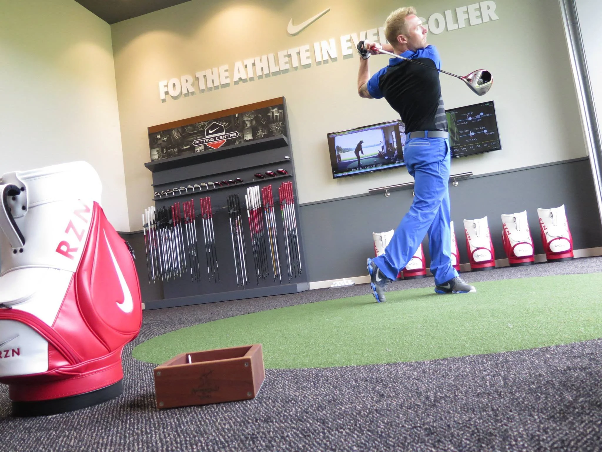 A person with blonde hair wearing a blue and black athletic shirt and blue pants practicing golf in an indoor golf simulation room. The room has a wall decorated with the Nike logo and a quote, a display of golf clubs, and a row of white golf bags with red accents. A screen on the wall shows a golf swing analysis.