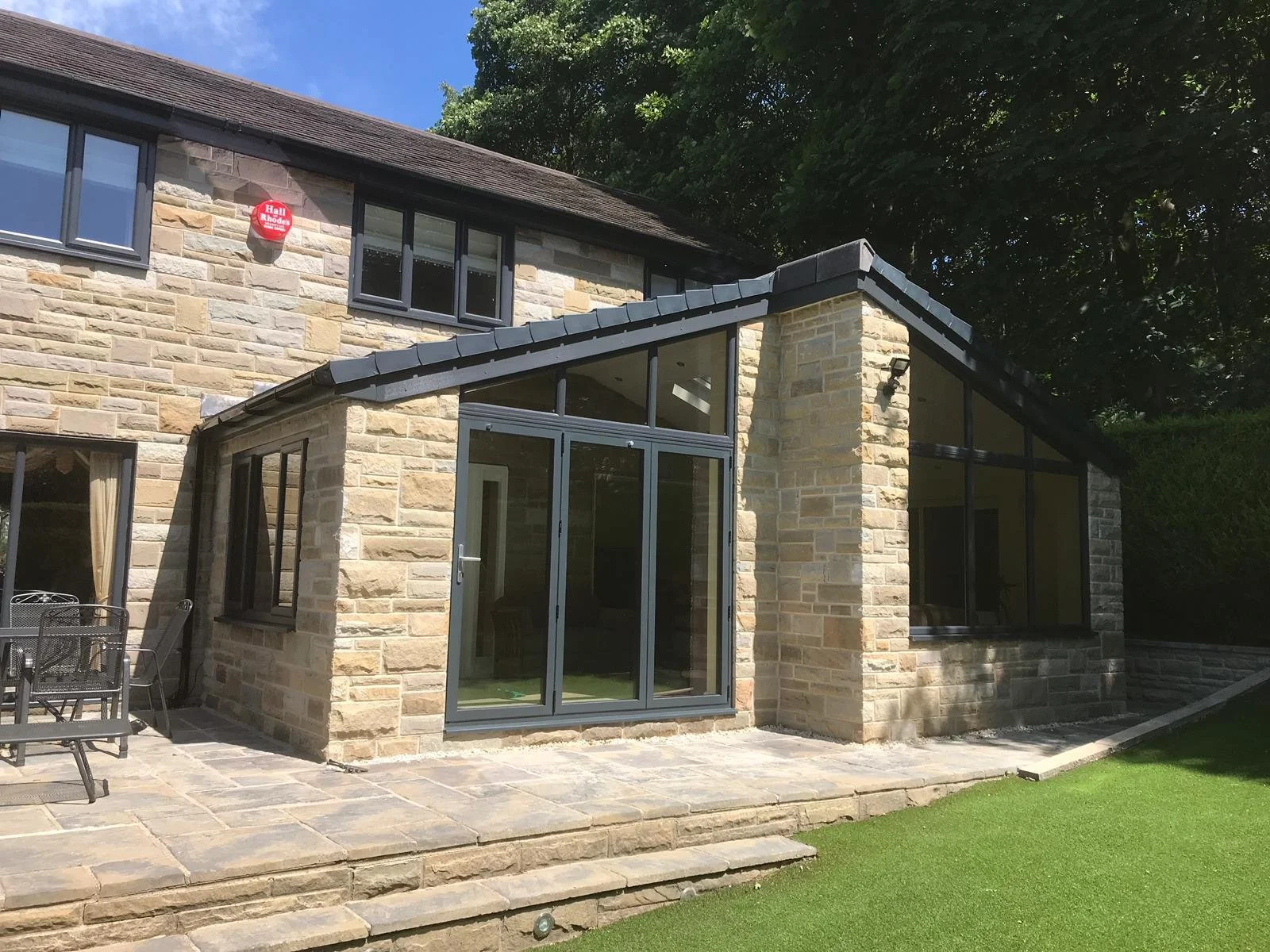 A modern stone house with large windows and a patio area with outdoor furniture, surrounded by a well-maintained green lawn and trees.