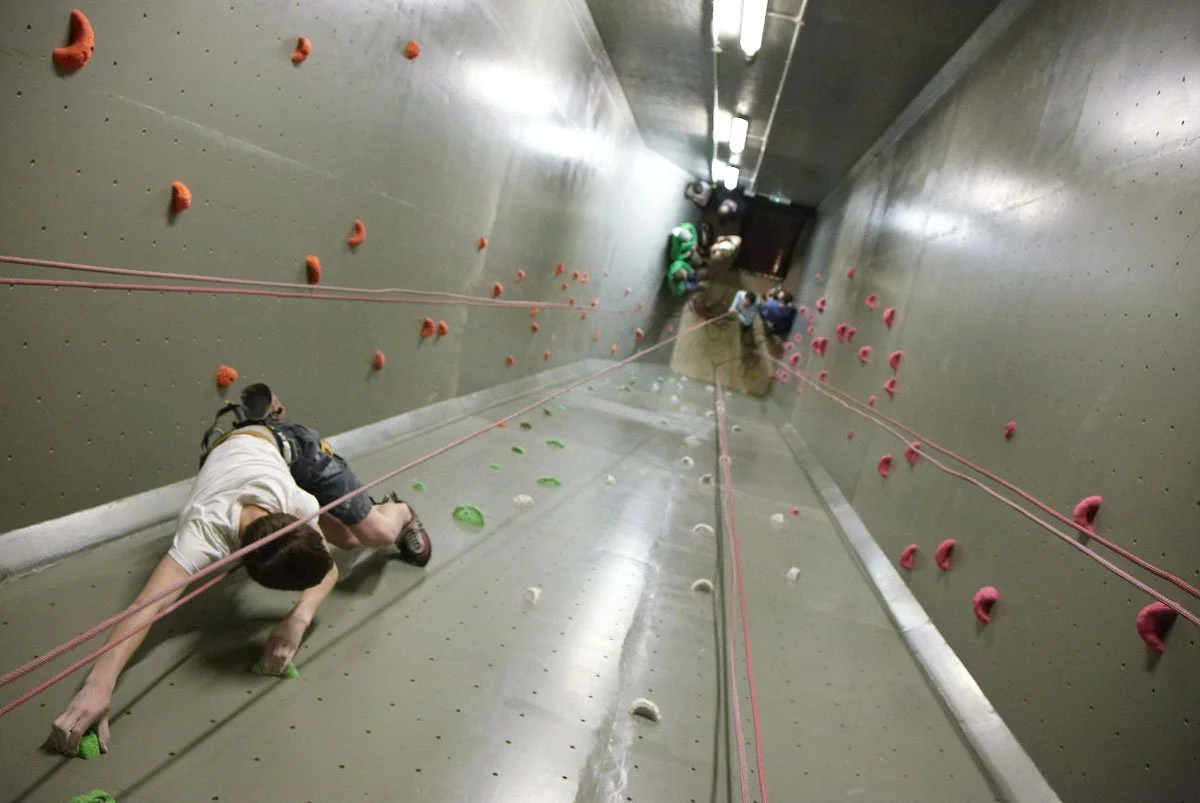 People climbing an indoor rock wall with various grips and a floor for safety.