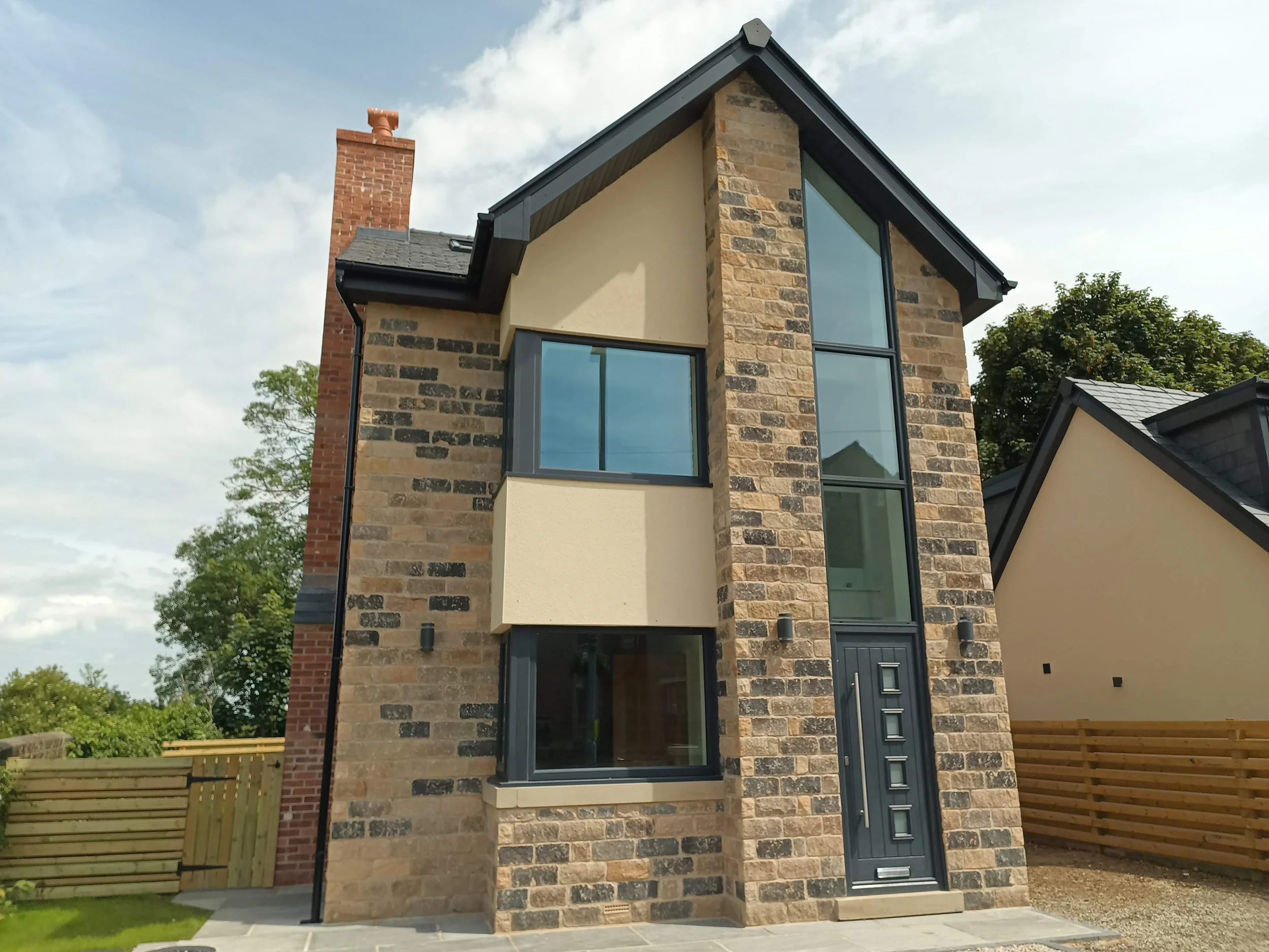 A modern brick house with large windows, a black front door, and a chimney, surrounded by a wooden fence and trees under a partly cloudy sky.
