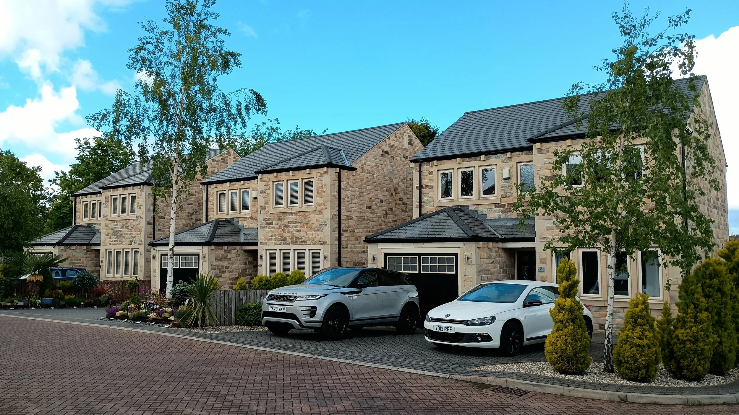 View of modern residential houses made of stone, parked cars in front, and landscaped gardens with trees and plants under a blue sky.