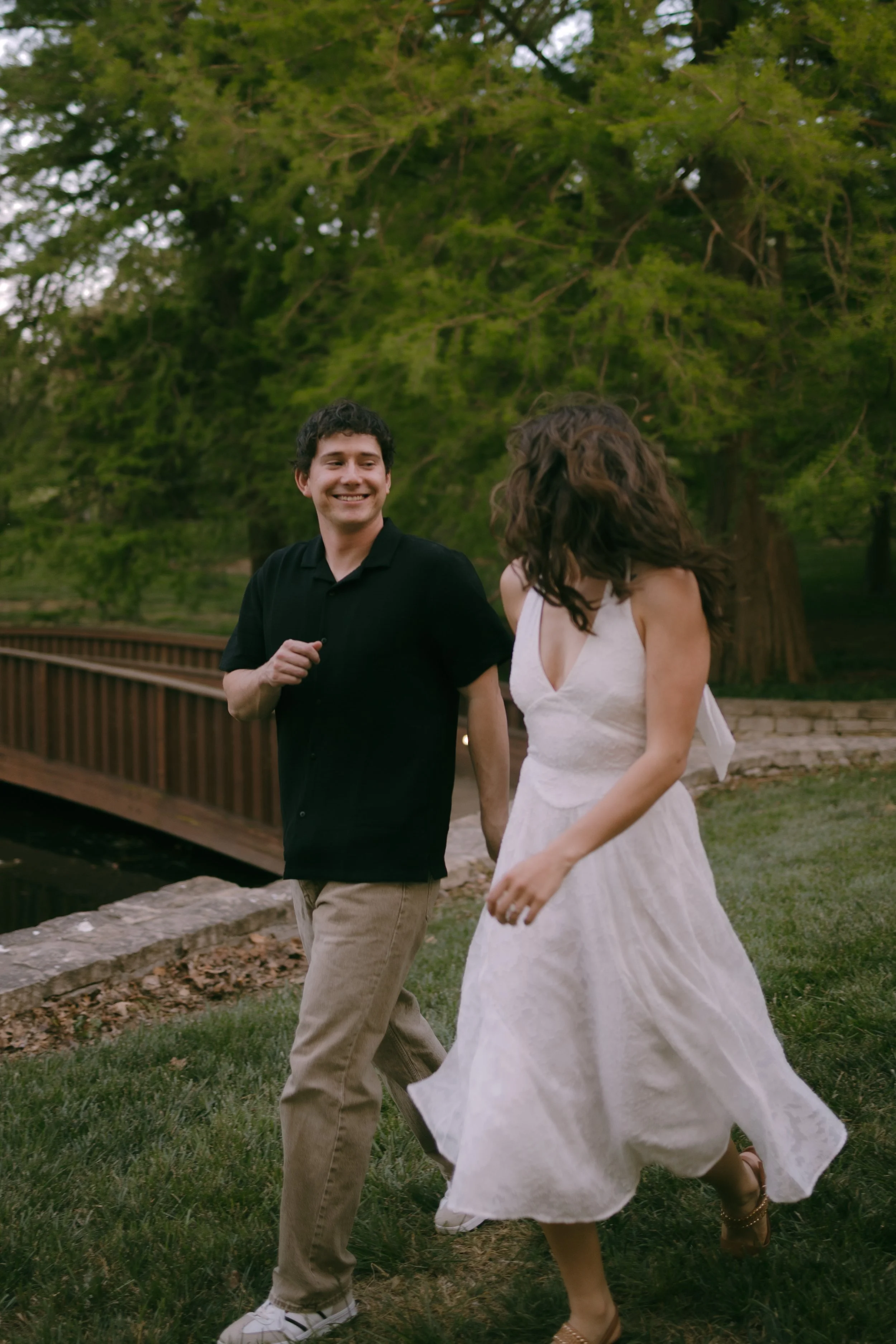 A young couple, smiling and holding hands, walking outside in a park with green grass, trees, and a wooden bridge in the background.