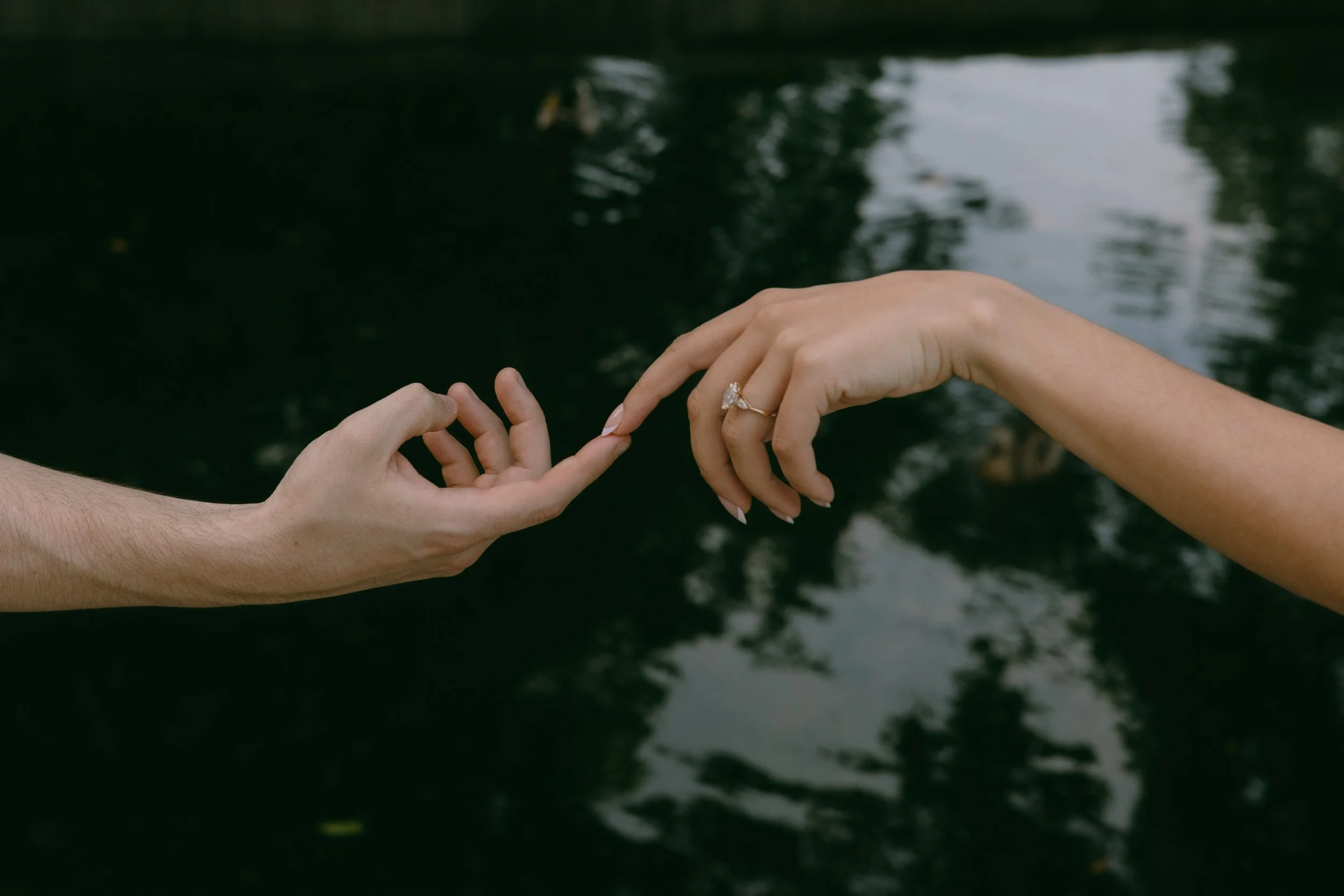 Two hands reaching towards each other above a body of water, with one hand wearing an engagement ring.