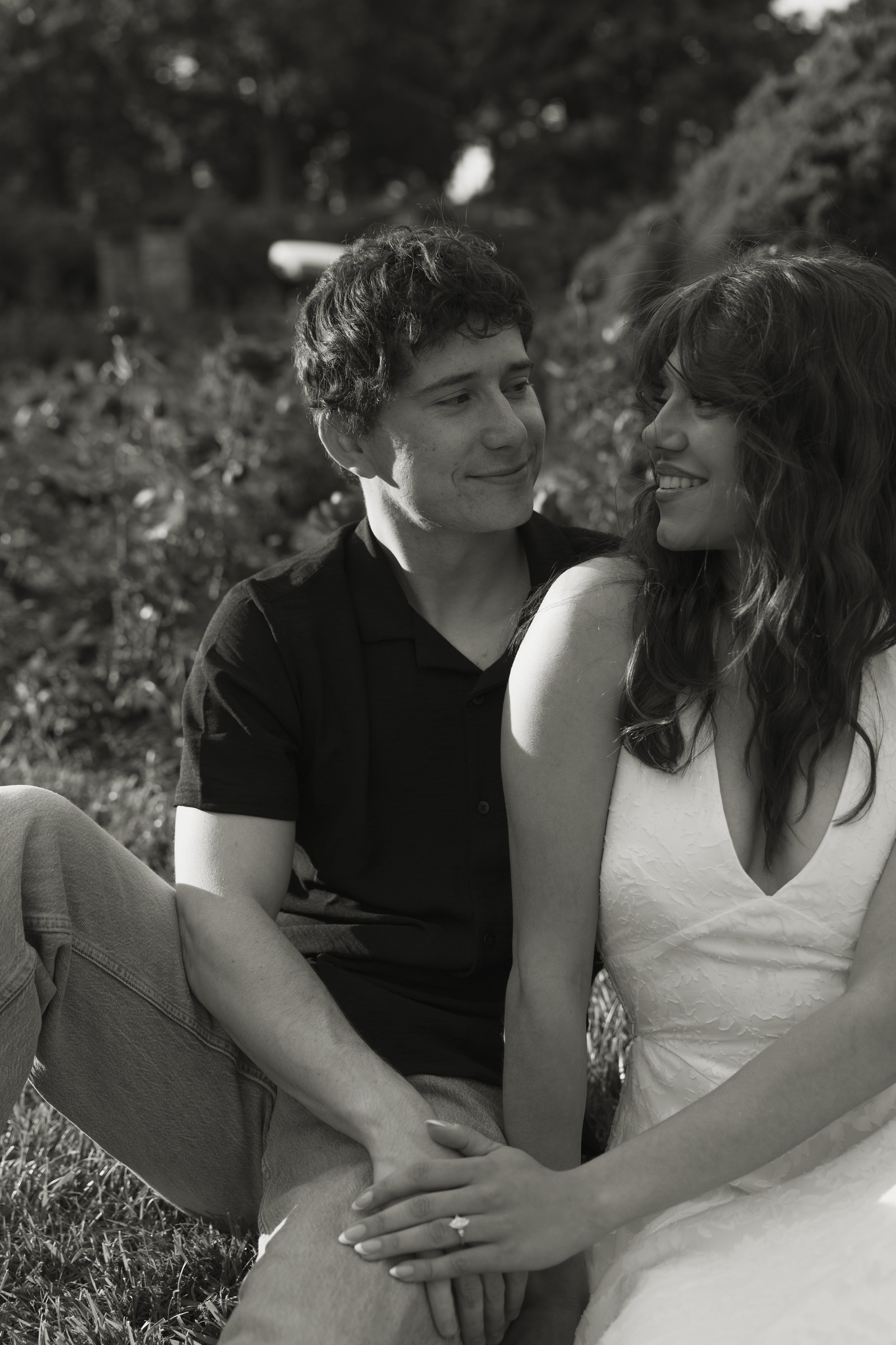 A black-and-white photo of a young couple sitting on the grass outdoors, smiling at each other closely. The man has short curly hair and is wearing a black shirt, while the woman has long curly hair and is wearing a white dress. They are gently holding each other's hands.